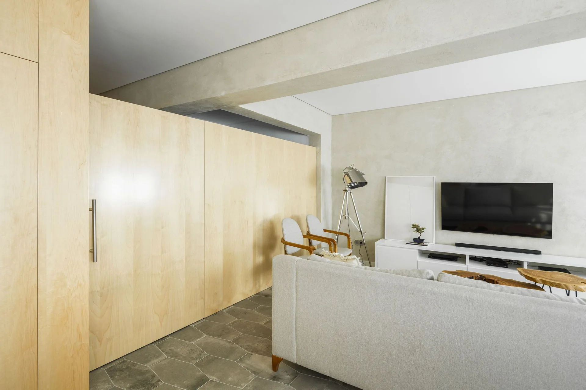 A modern living room featuring a light wood paneled wall partition, an exposed concrete ceiling beam, grey hexagonal floor tiles, and a white TV console.