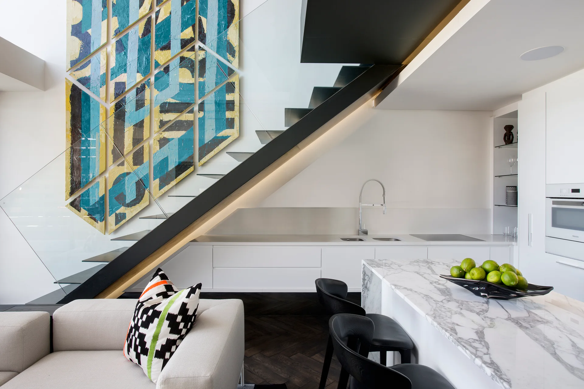 A modern kitchen with a white marble island, black bar stools, a floating staircase with glass railings, and a large blue and yellow abstract painting.