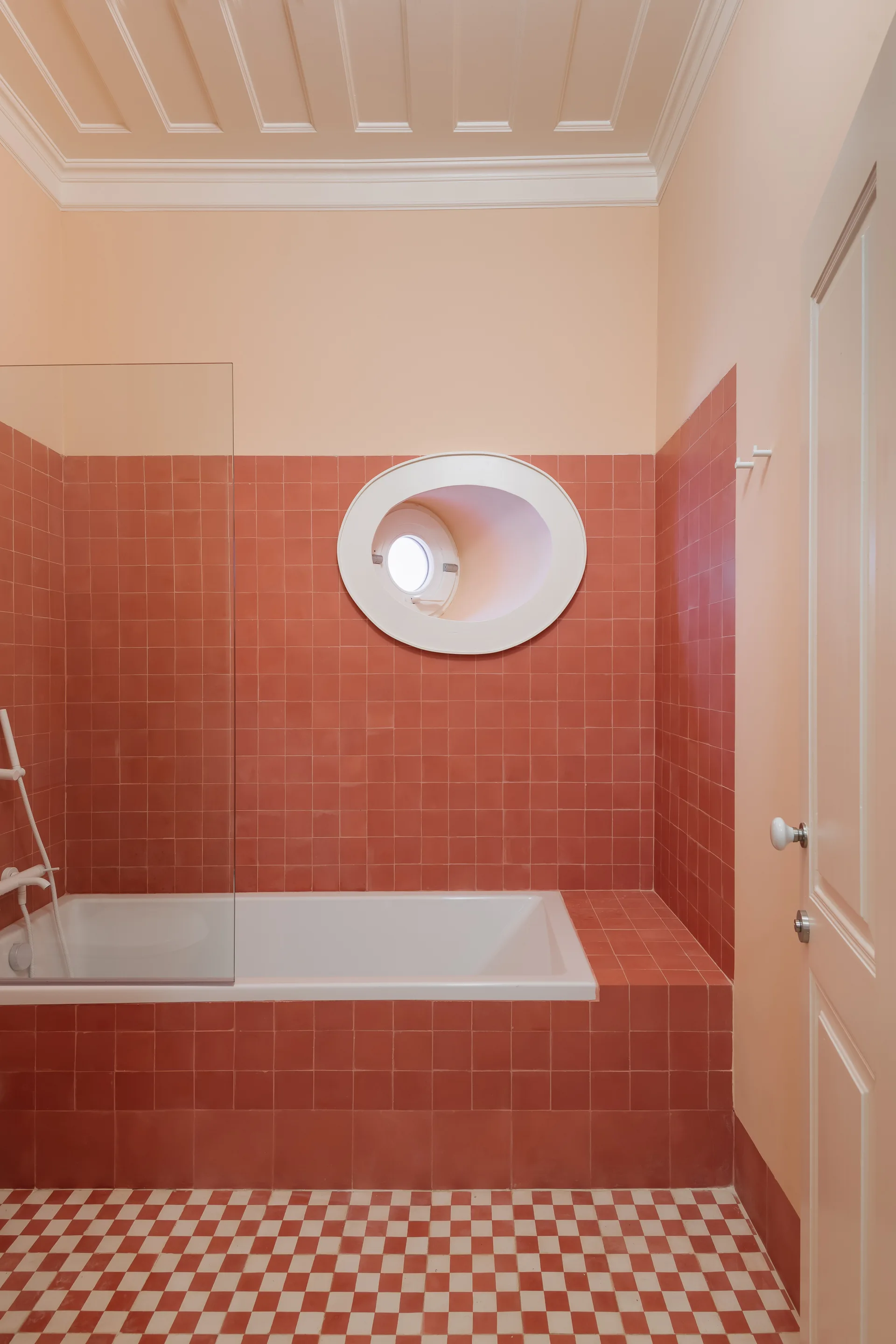 A bathroom featuring a built-in tub with terracotta tiled walls, a small oval porthole window, and a red and white checkerboard floor.