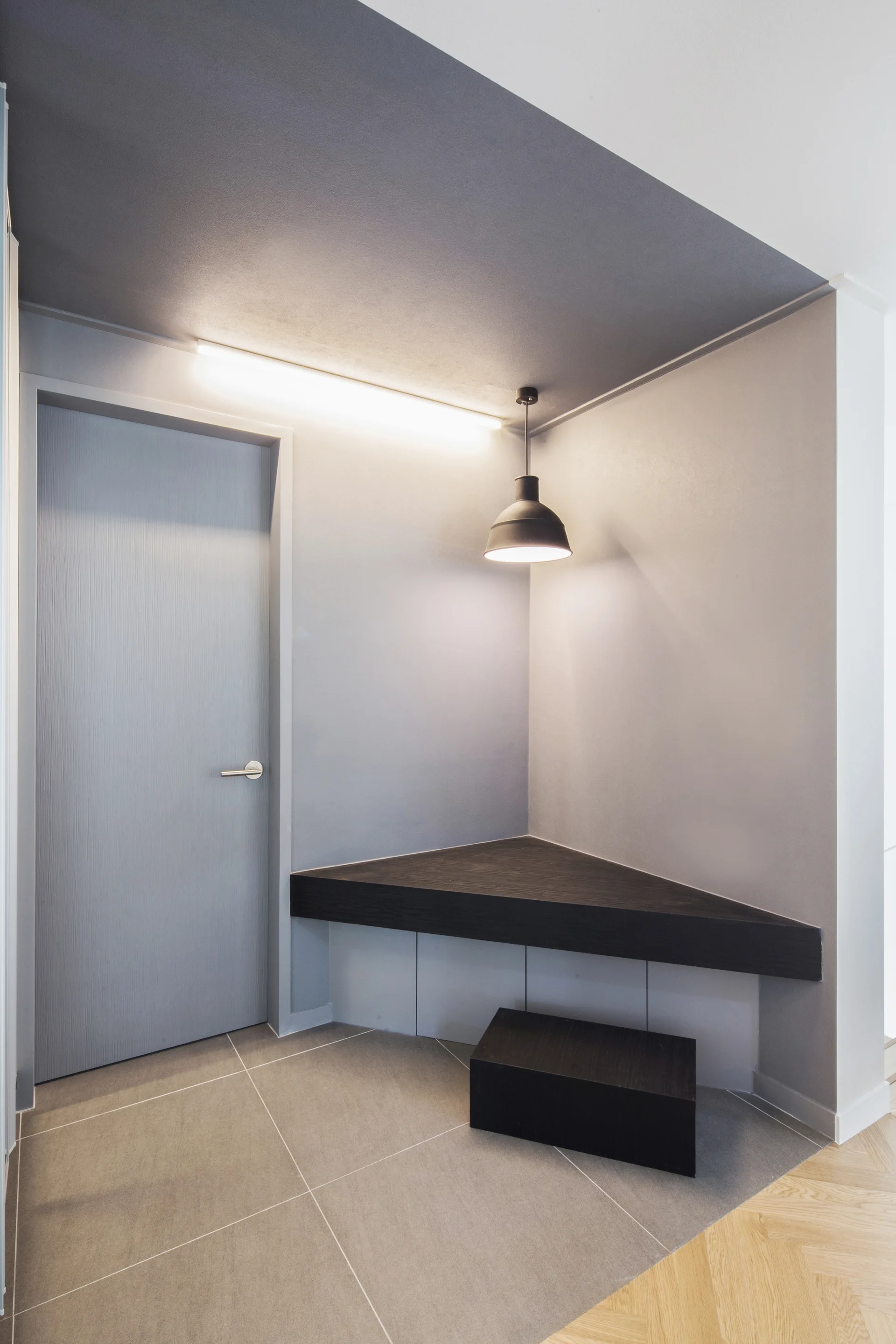 A minimalist grey entryway with a flush door, a floating dark wood corner bench, a matching block stool, and a black pendant light.