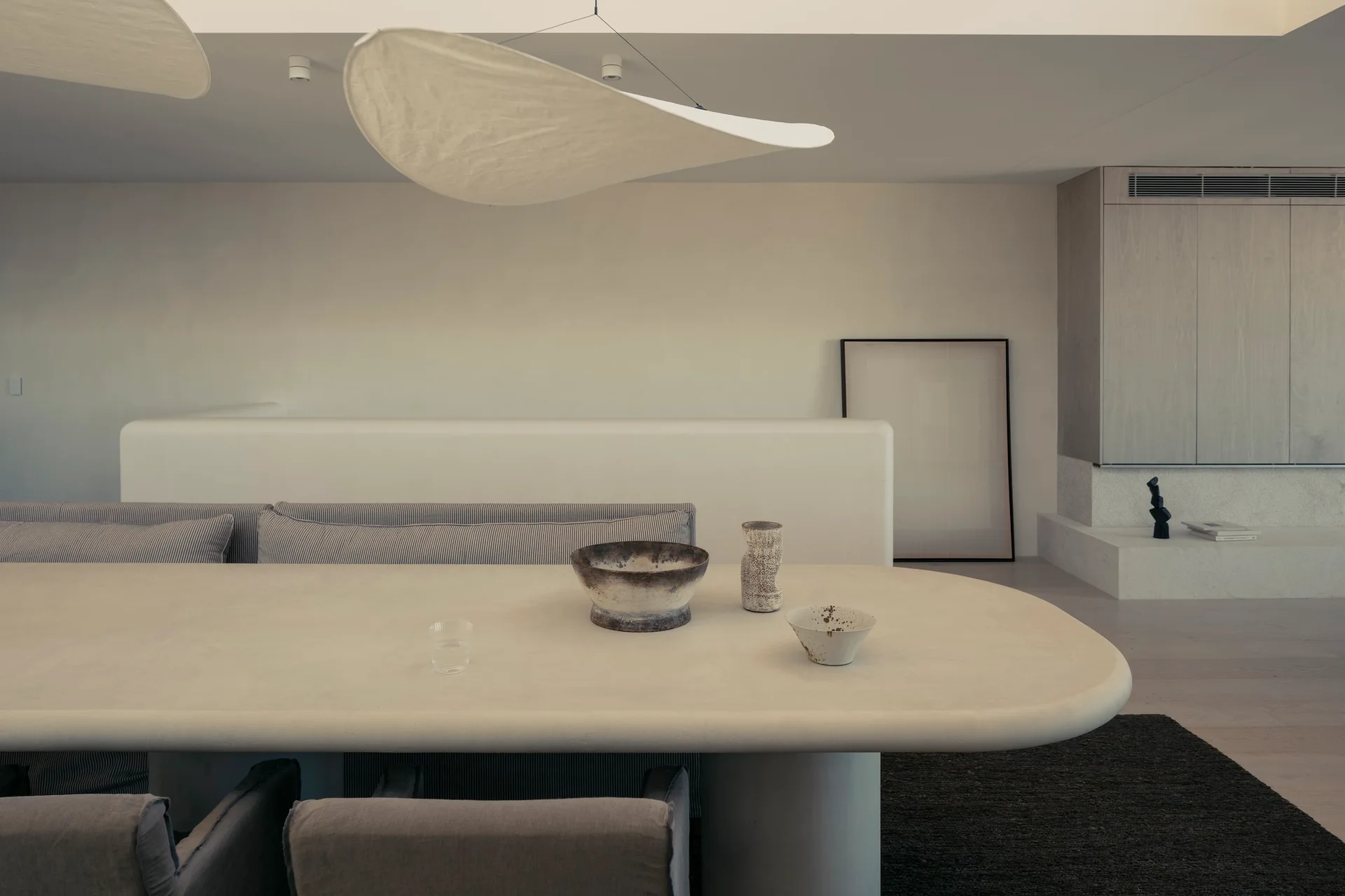 A minimalist dining area featuring a large cream plaster dining table with striped bench seating and a floating organic fabric pendant light.