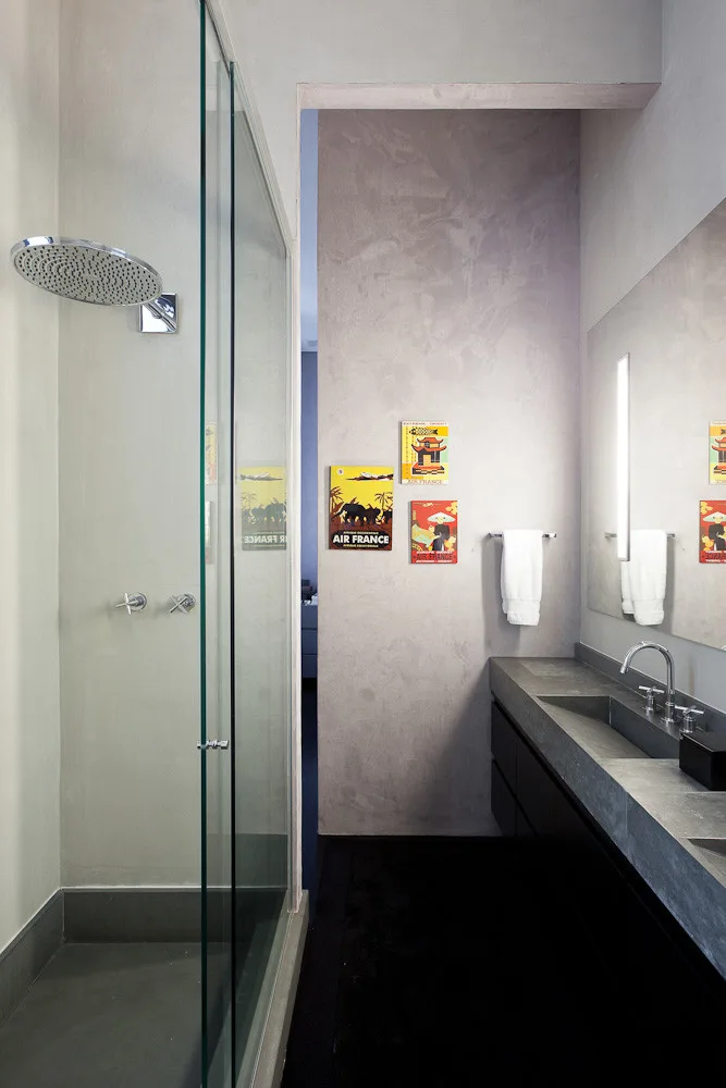A contemporary bathroom with a glass-enclosed shower, a concrete trough sink vanity, textured plaster walls, and colorful vintage wall posters.