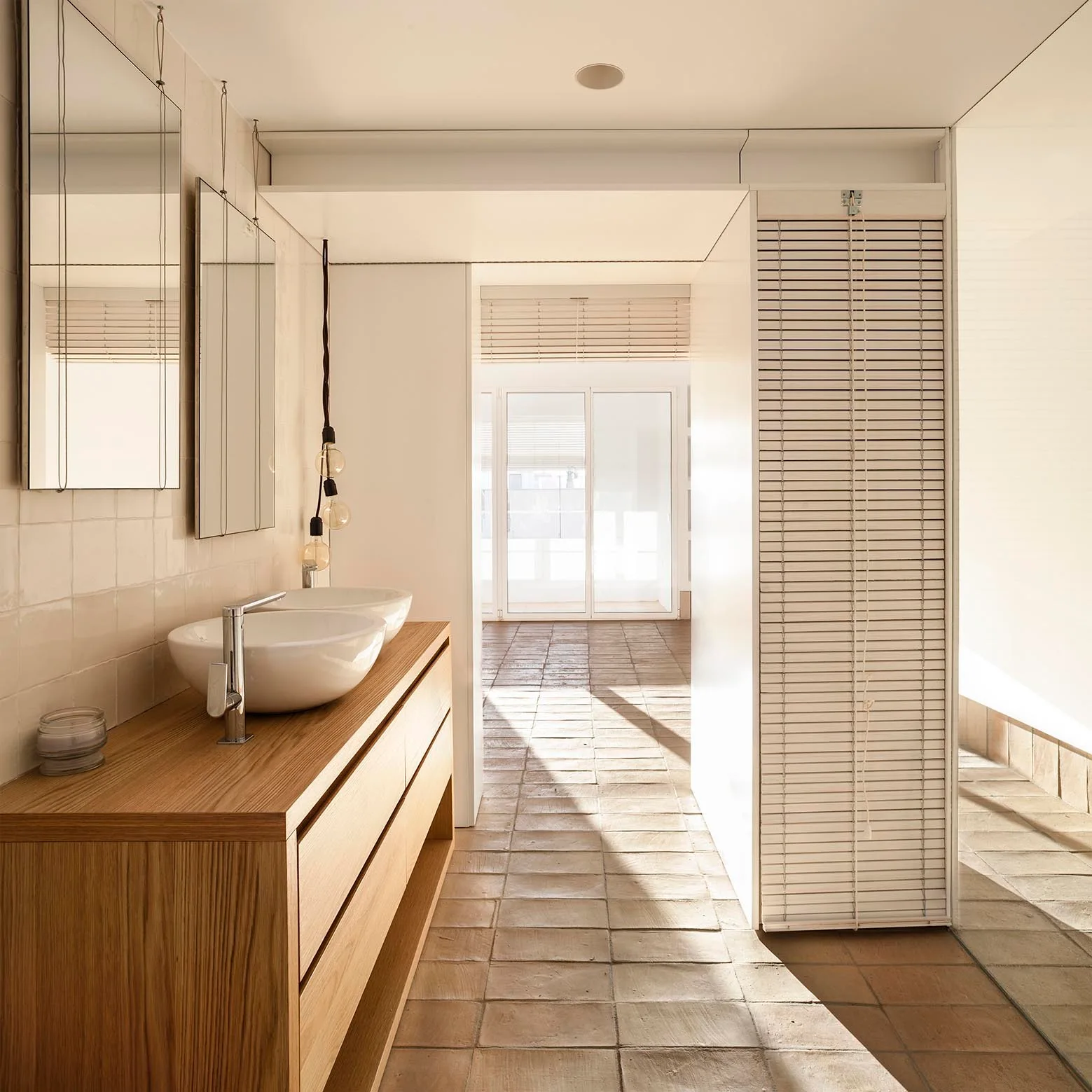 A modern bathroom features a light wood double vanity with white vessel sinks, square cream wall tiles, and matte terracotta floor tiles.