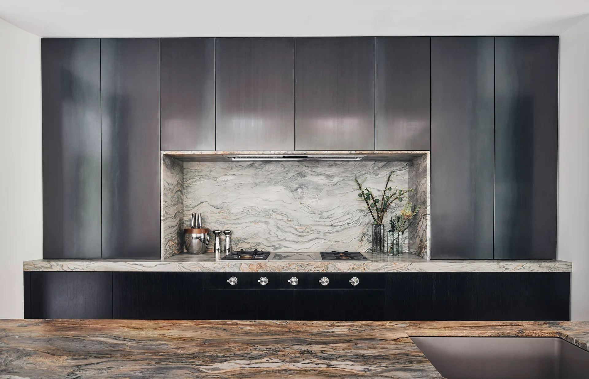 A modern kitchen featuring dark metallic-finish upper cabinets, black base cabinets, and a dramatic veined stone backsplash and countertop.