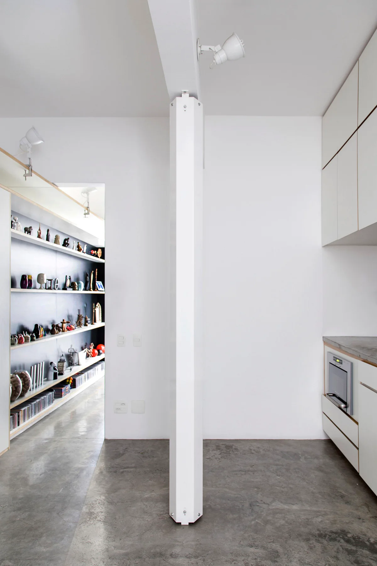 A minimalist kitchen with white cabinetry and a central white column stands on polished concrete flooring next to an open doorway showing a shelving unit.
