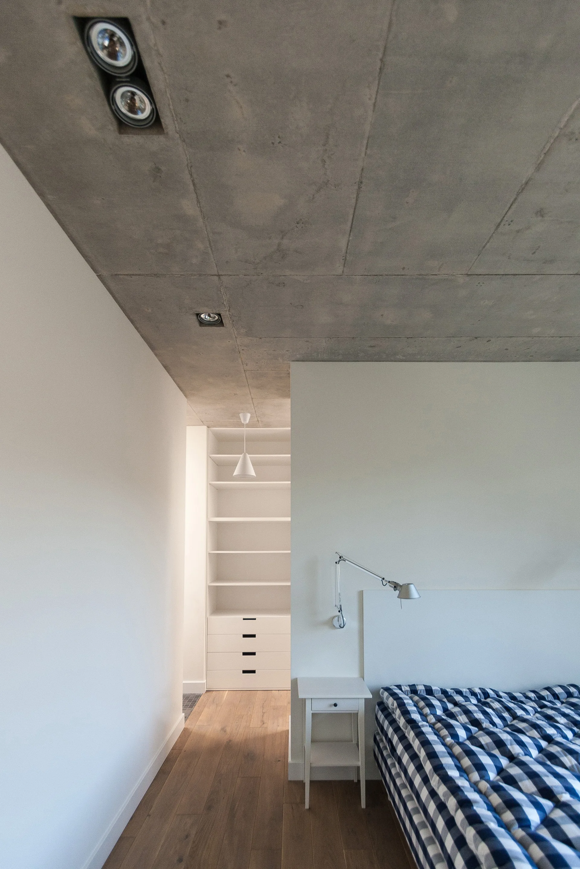 A minimalist bedroom featuring an exposed concrete ceiling, a white wall-mounted nightstand, and a bed with a blue checkered quilt.