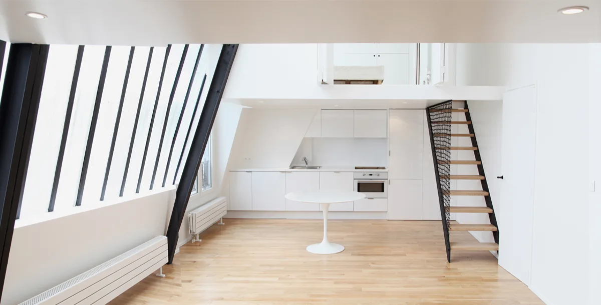 A minimalist loft apartment features a white kitchen under a mezzanine, a central white pedestal table, light wood floors, and a slanted window wall.