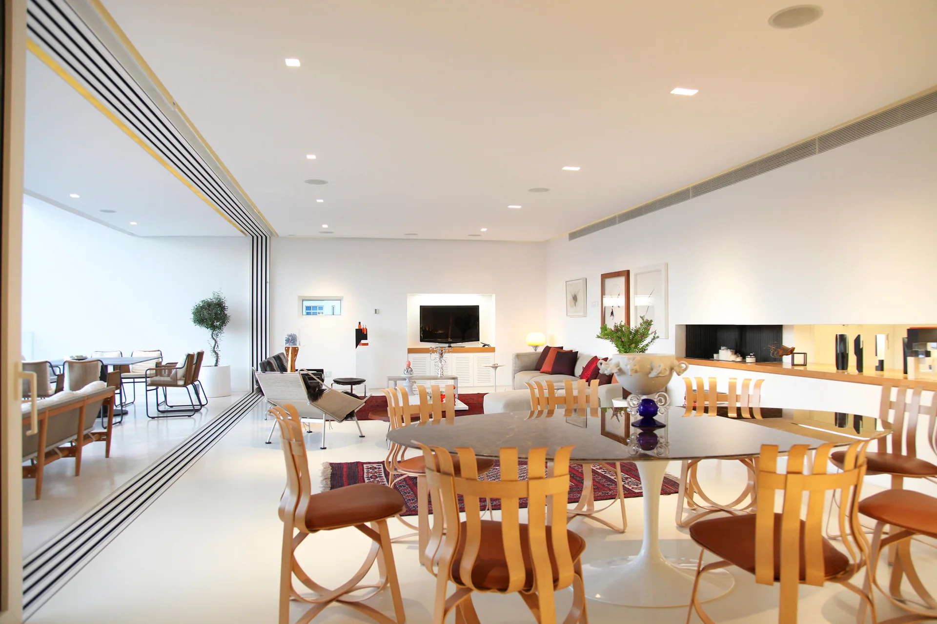 A large white-walled dining and living space featuring a round table with curved wooden chairs and a sliding glass wall.