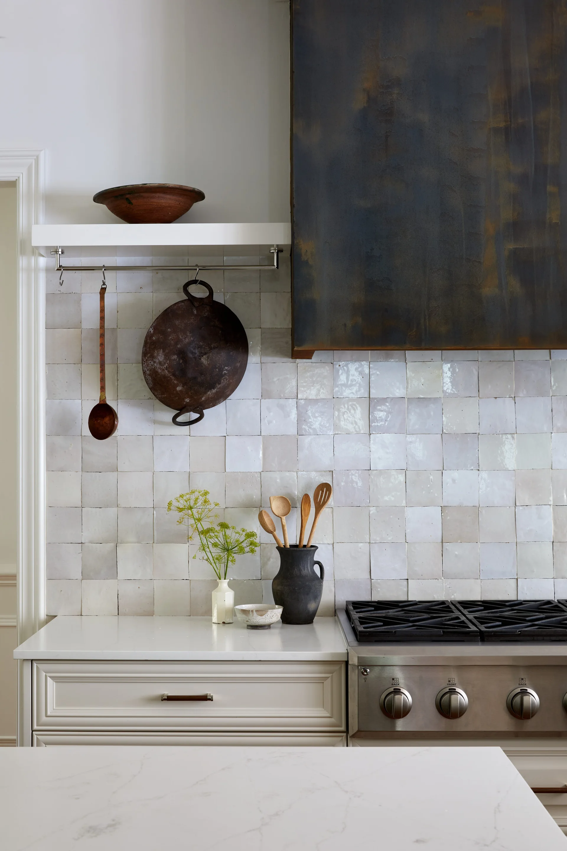 A kitchen stove features a dark patinated metal range hood mounted over a glazed white square tile backsplash with a white floating shelf.