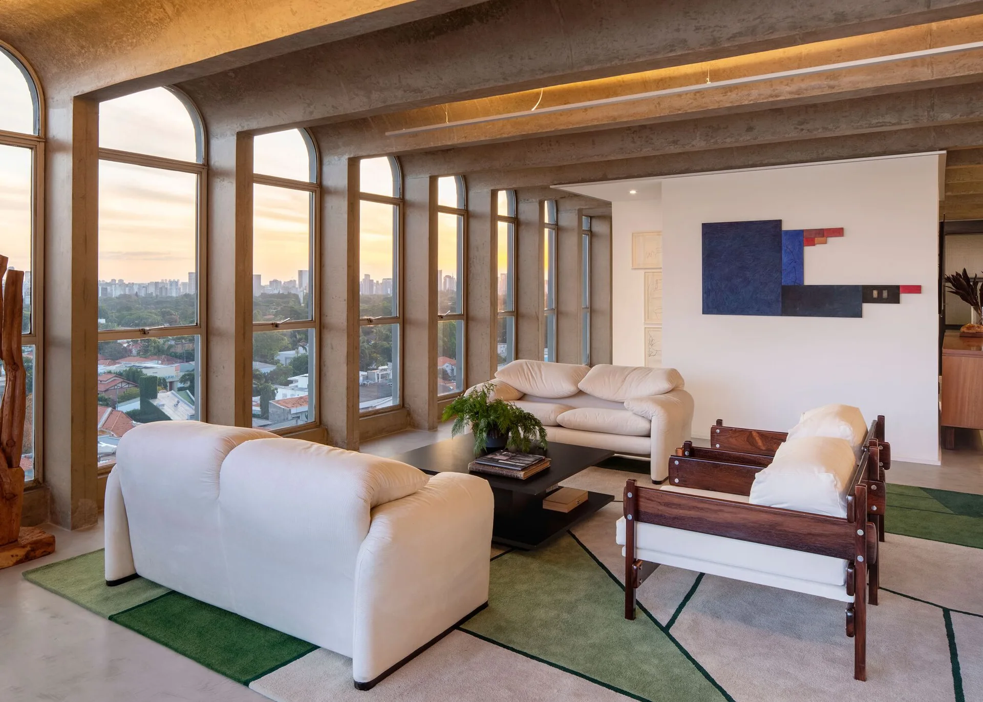 A living room with white sofas, wooden lounge chairs, and a geometric rug is framed by arched concrete windows and exposed ceiling beams.