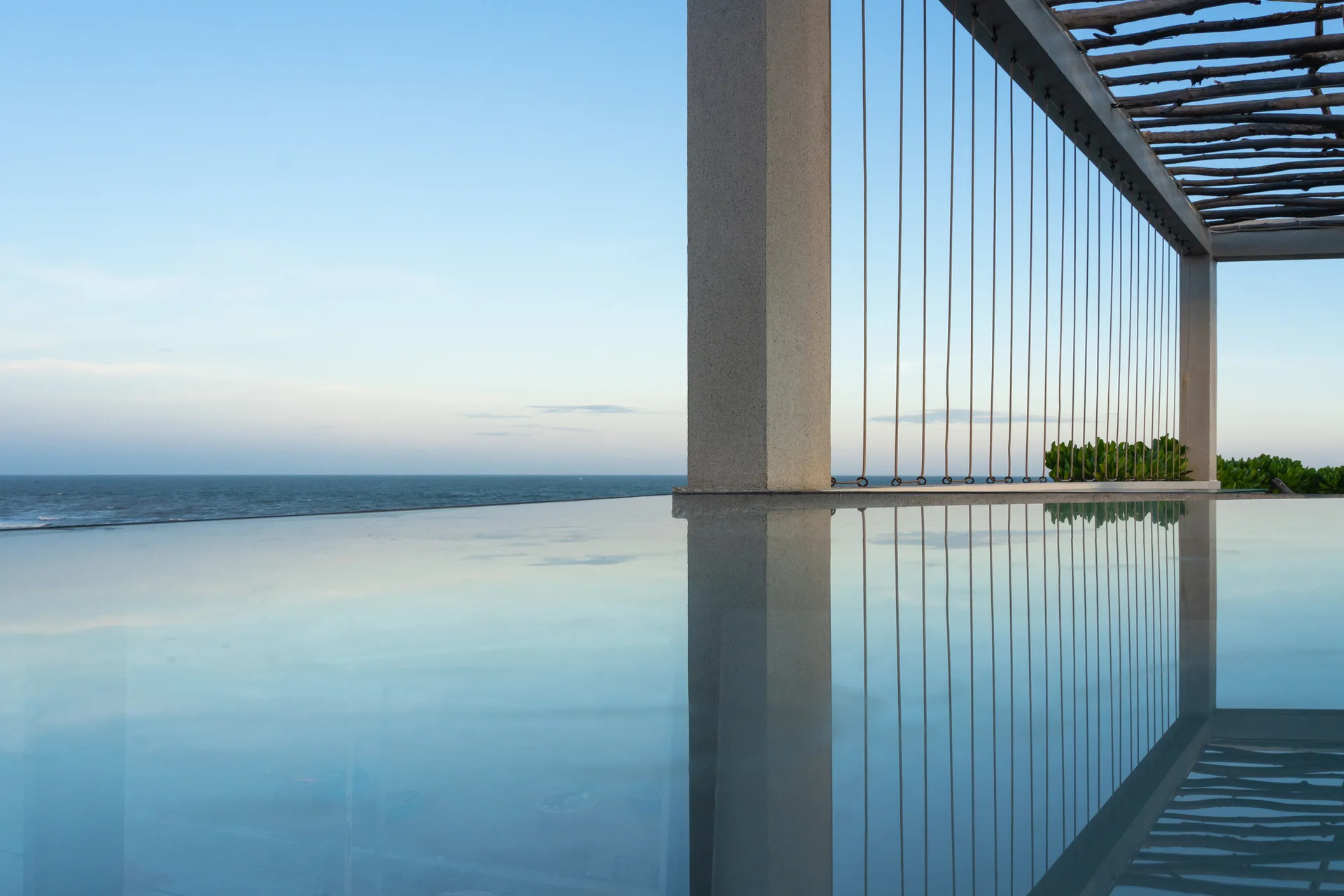 An outdoor infinity-edge pool reflects a stone-like column and a vertical cable railing with an ocean view in the background.