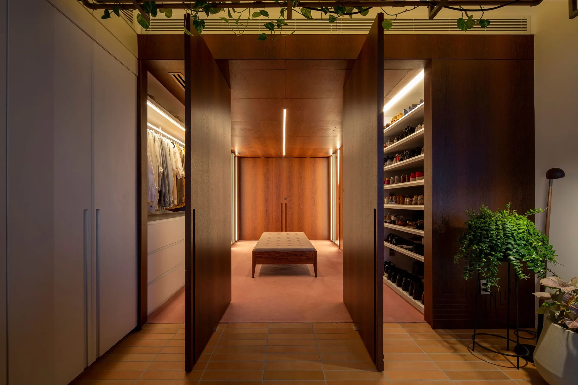 A walk-in closet with dark wood paneling, integrated vertical lighting, a tufted bench on a pink carpet, and open shelves containing clothes and shoes.