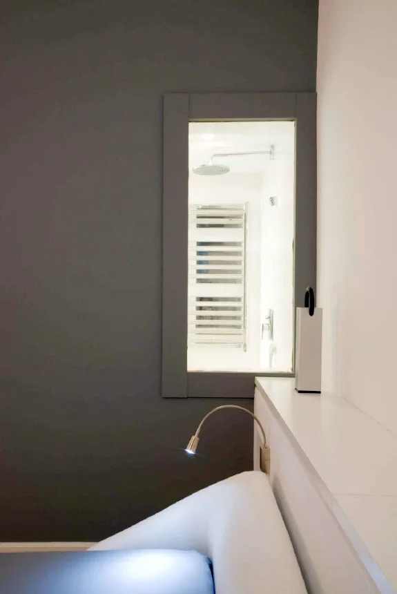 A dark grey bedroom wall features a small square interior window looking into a white bathroom with a visible shower head.