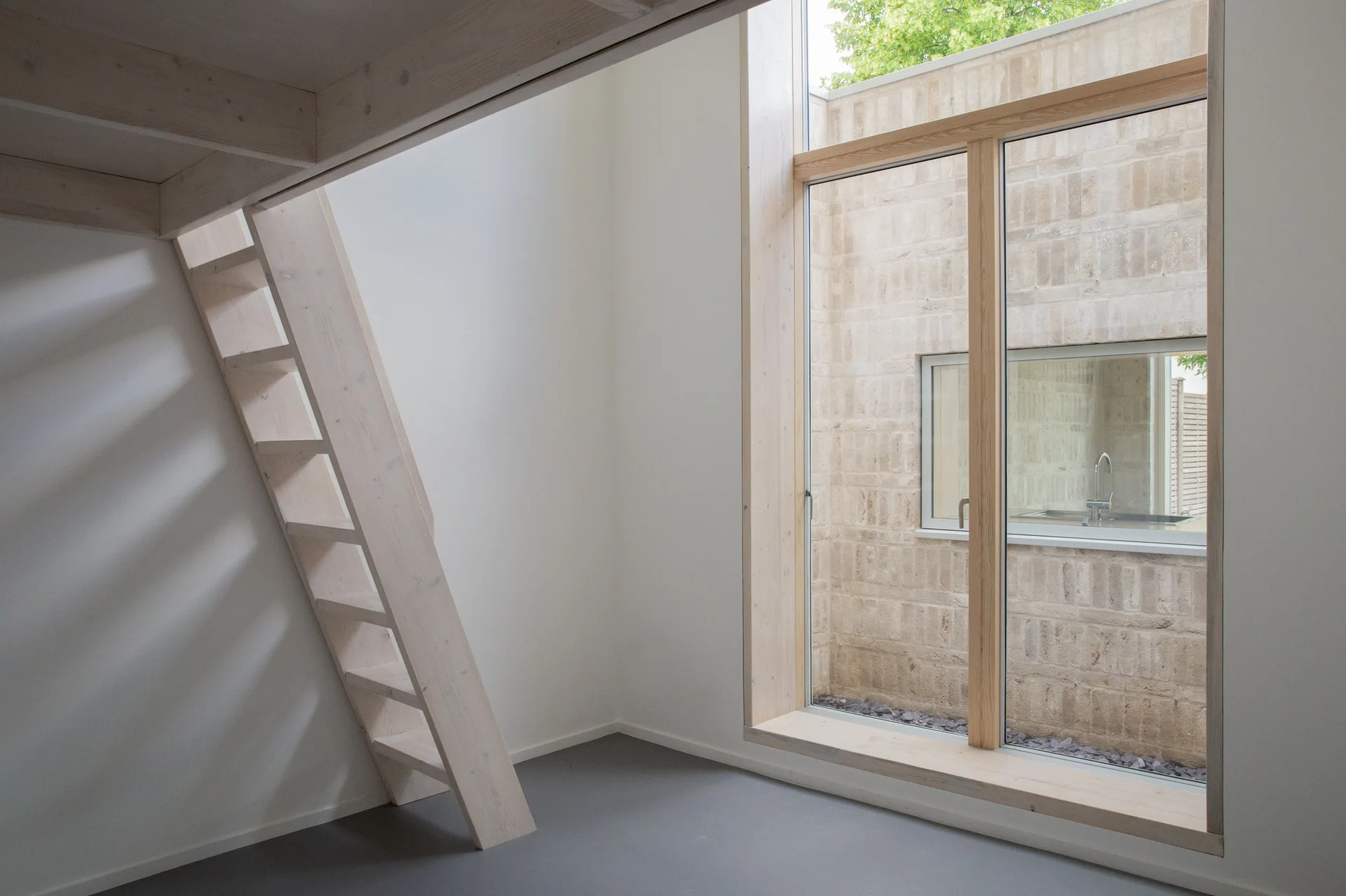 A minimalist room features a light wood ladder leading to a loft, a large wood-framed window, and a view of a brick courtyard.