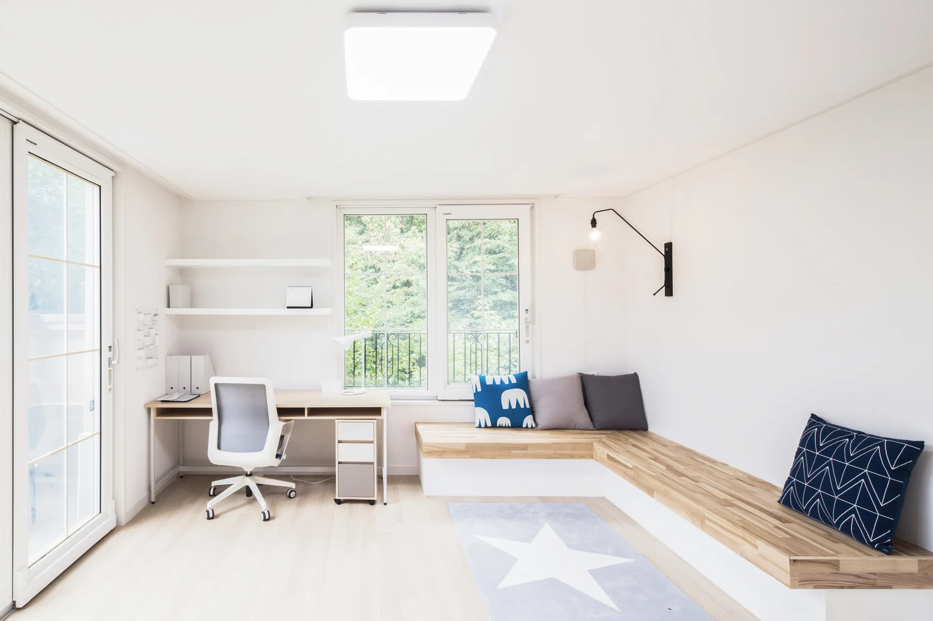 A bright home office features a light wood desk, a grey-and-white task chair, and a low L-shaped built-in wooden bench beneath a window.