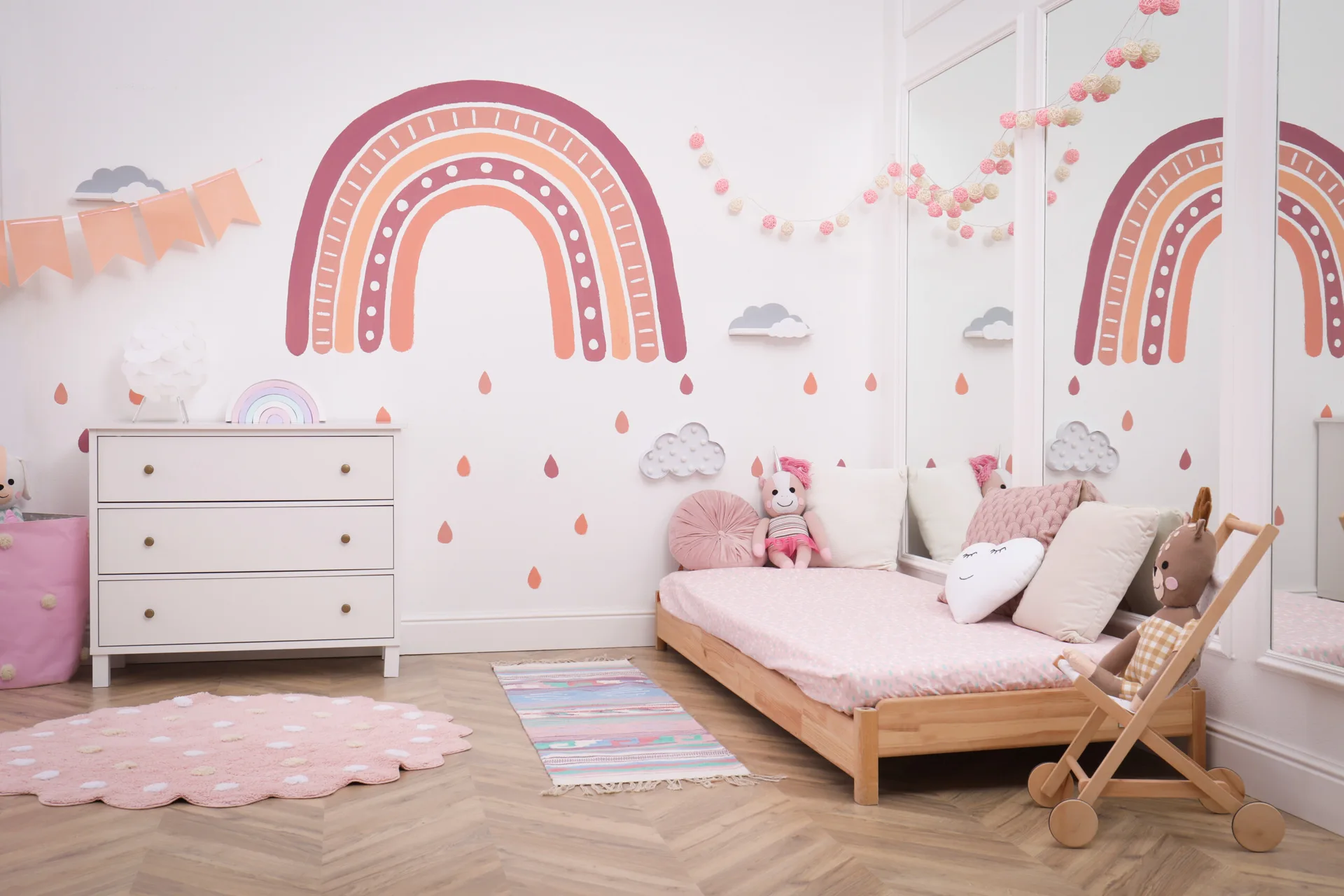 A children's bedroom features a low wooden bed, a white dresser, and a large pink and peach rainbow mural on the white wall.