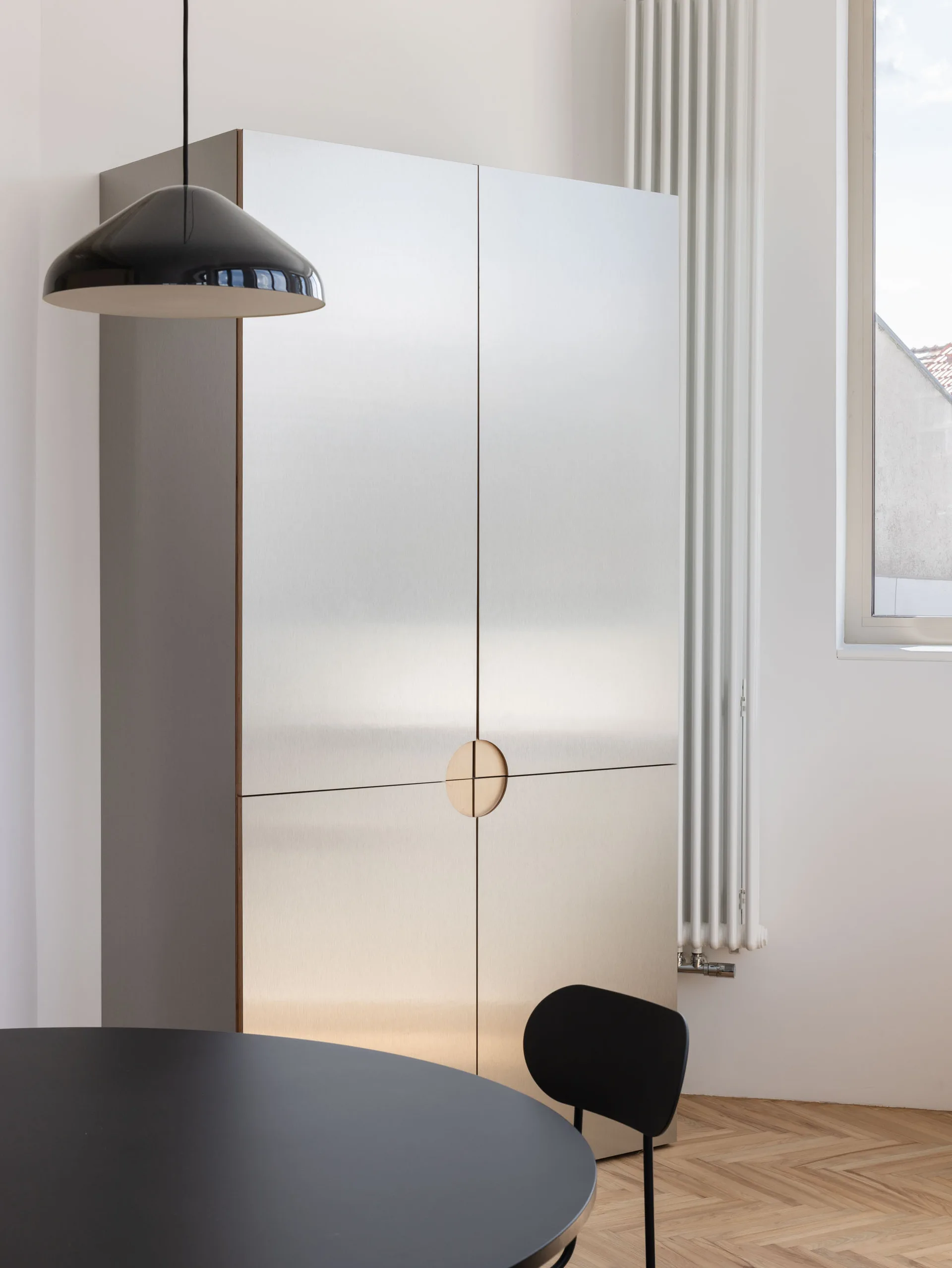 A large brushed metal storage cabinet with a circular recessed wooden handle stands in a minimalist room with herringbone parquet floors.