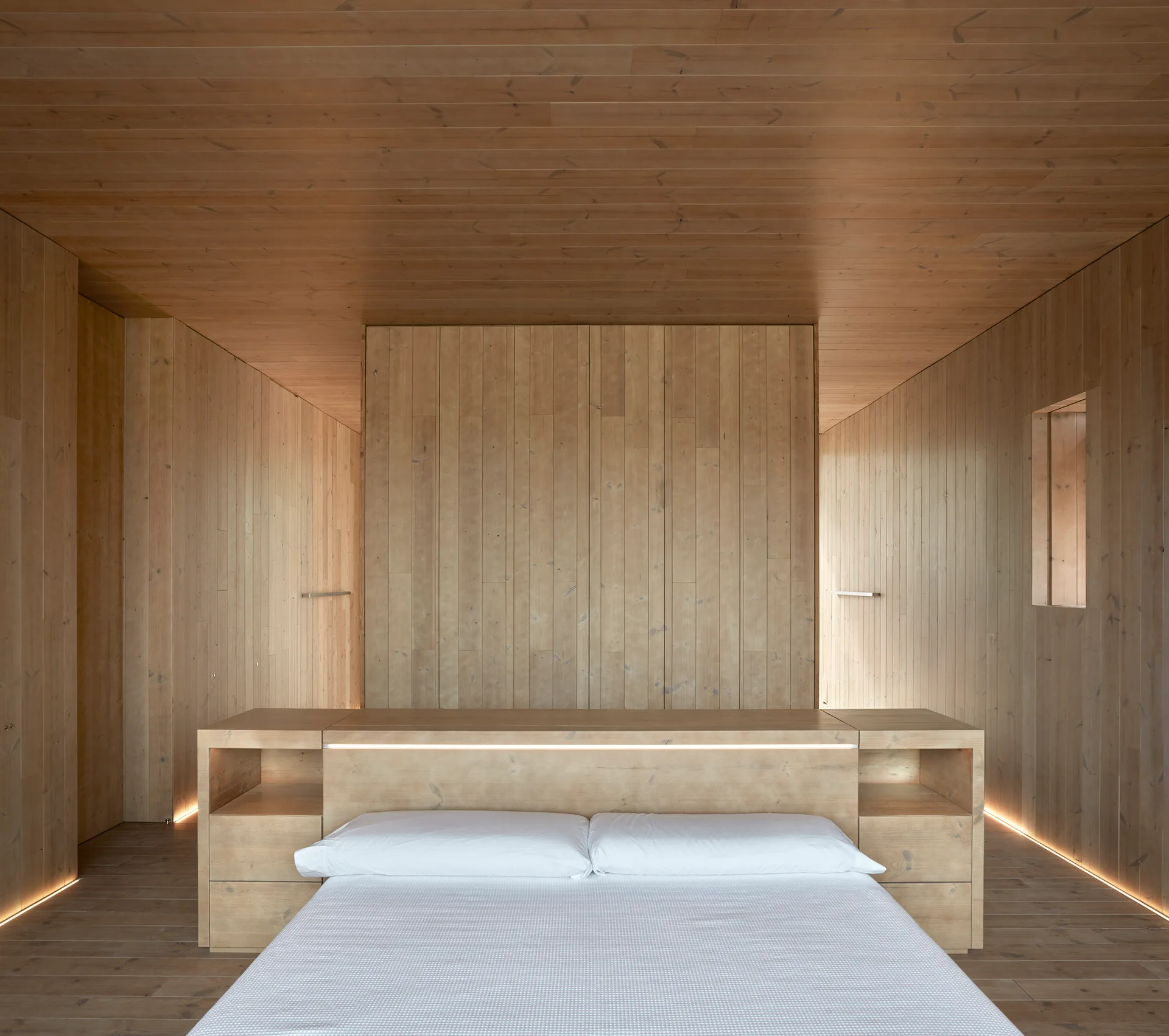 A minimalist bedroom features walls, ceiling, and floor clad in light-toned wood planks with an integrated wooden headboard and white bed linens.
