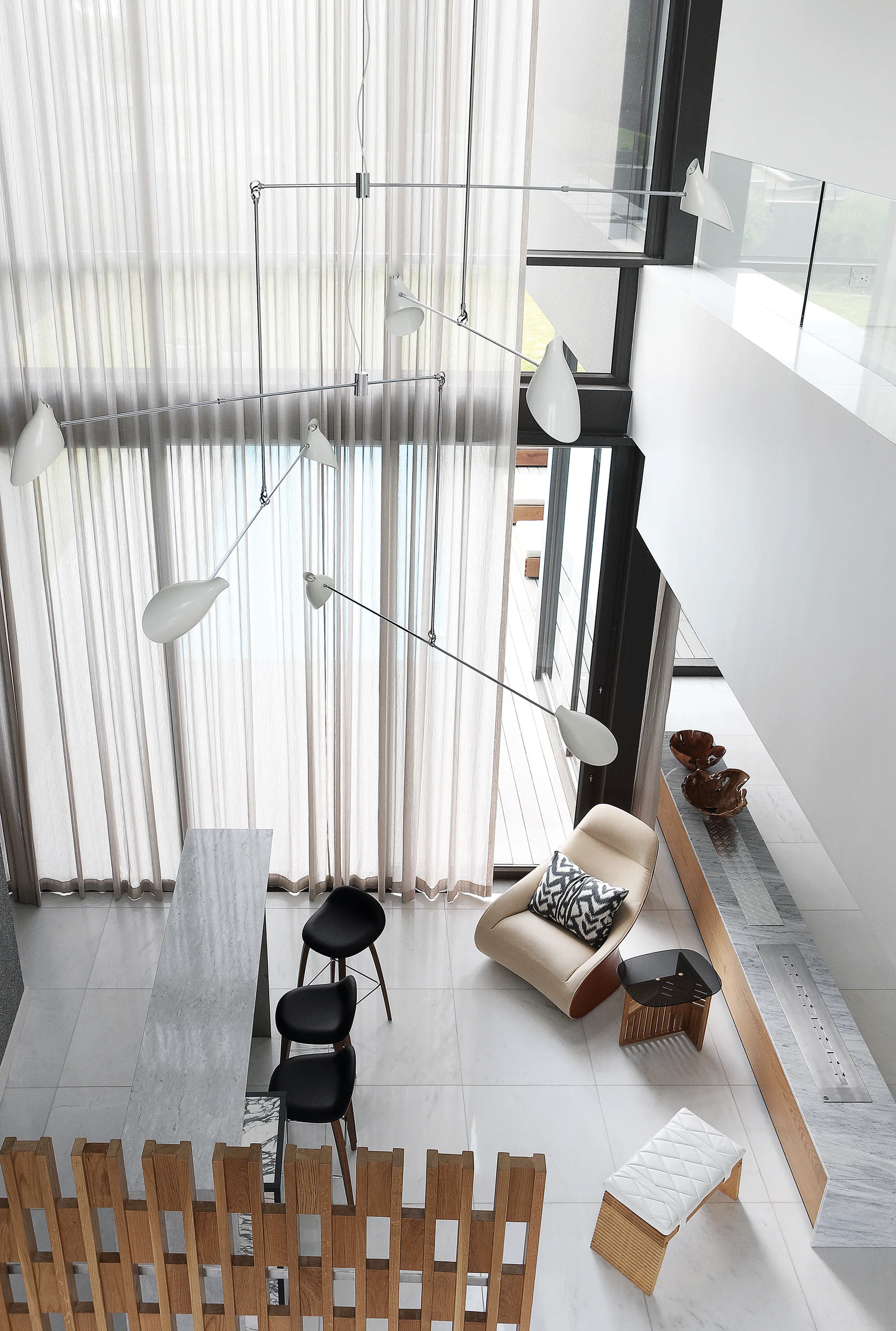 High-angle view of a double-height living room featuring a multi-arm white chandelier, sheer curtains, a stone counter, and a slatted wood partition.