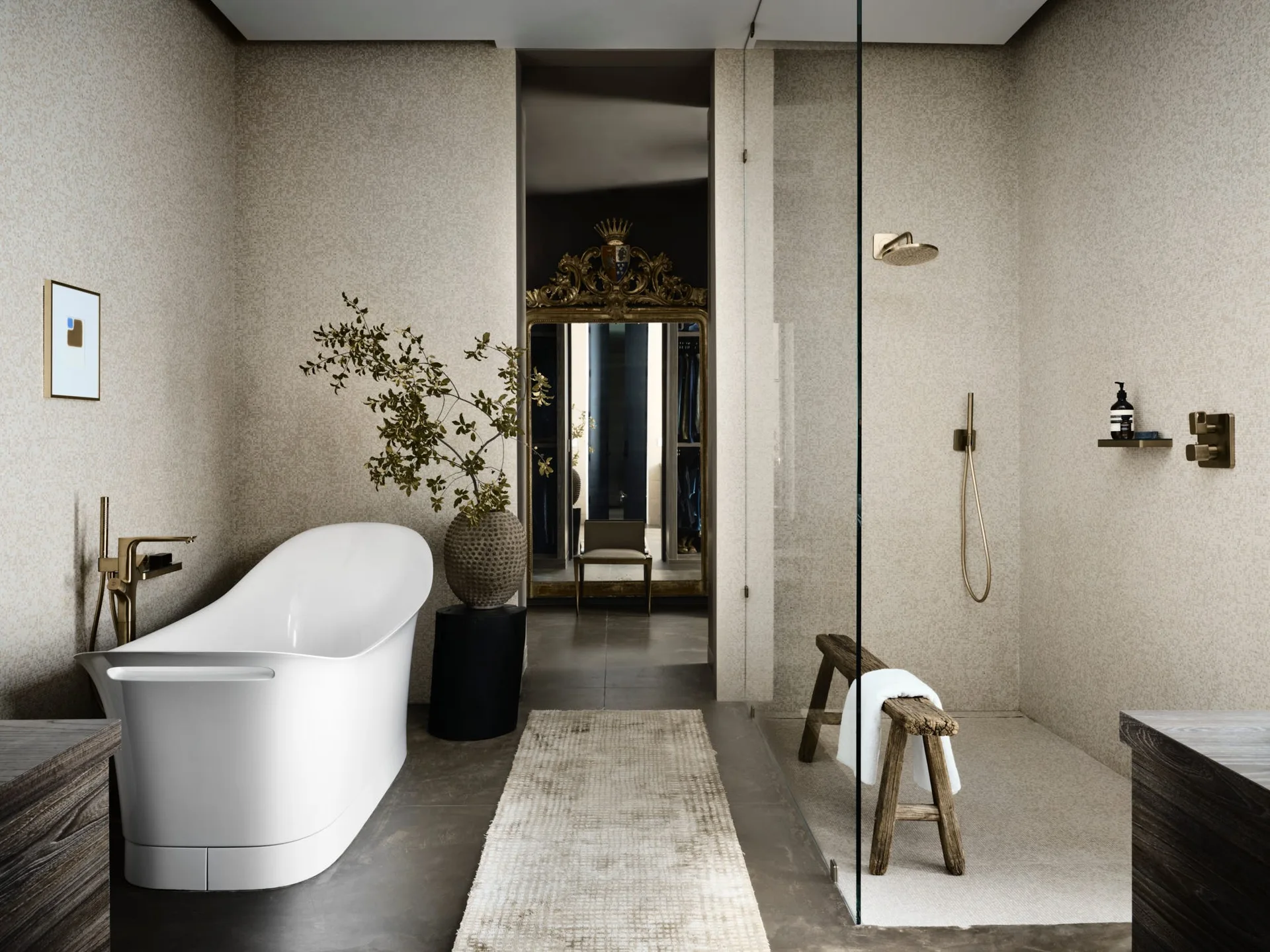 A modern bathroom features a white freestanding bathtub, textured beige walls, and a walk-in glass shower with a rustic wooden stool.
