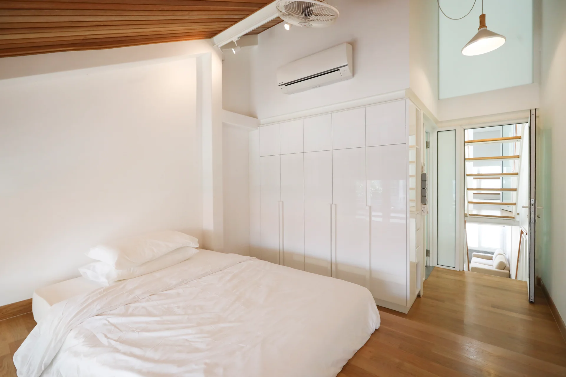 A minimalist bedroom features a low platform bed with white sheets, a large built-in white wardrobe, and a sloped wood plank ceiling.