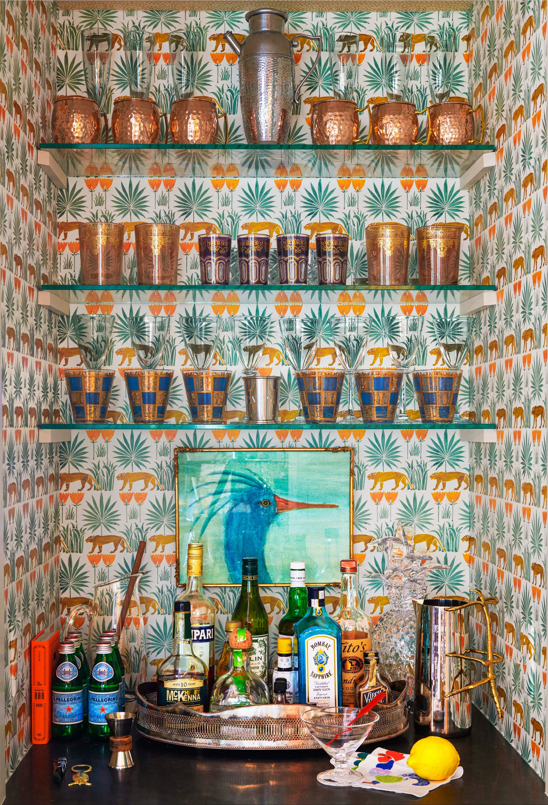 A built-in home bar niche features three glass shelves holding glassware and a cocktail shaker against green and orange animal-patterned wallpaper.