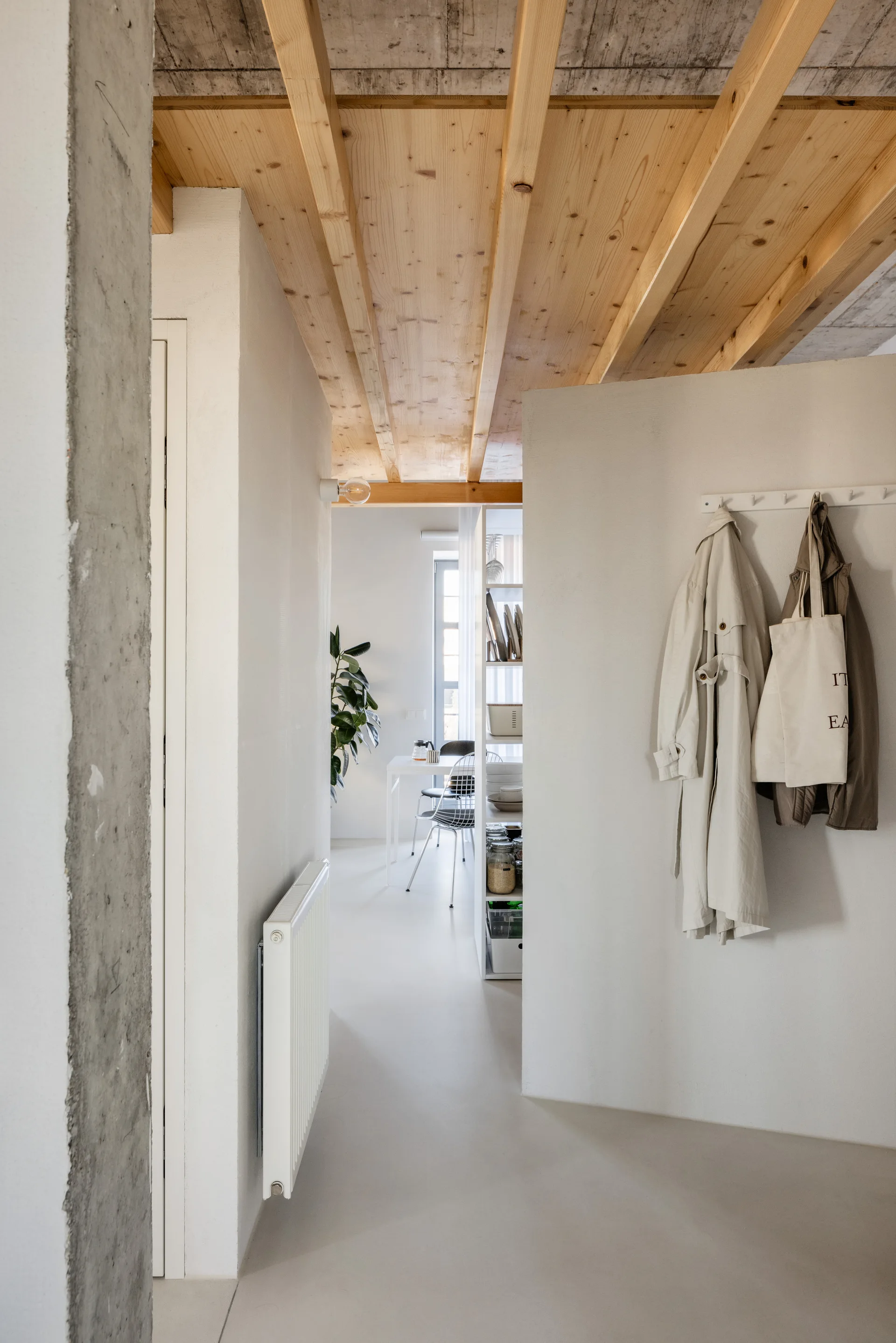 A minimalist white hallway featuring exposed wood ceiling beams, a wall-mounted coat rack, and a view into a bright dining area.