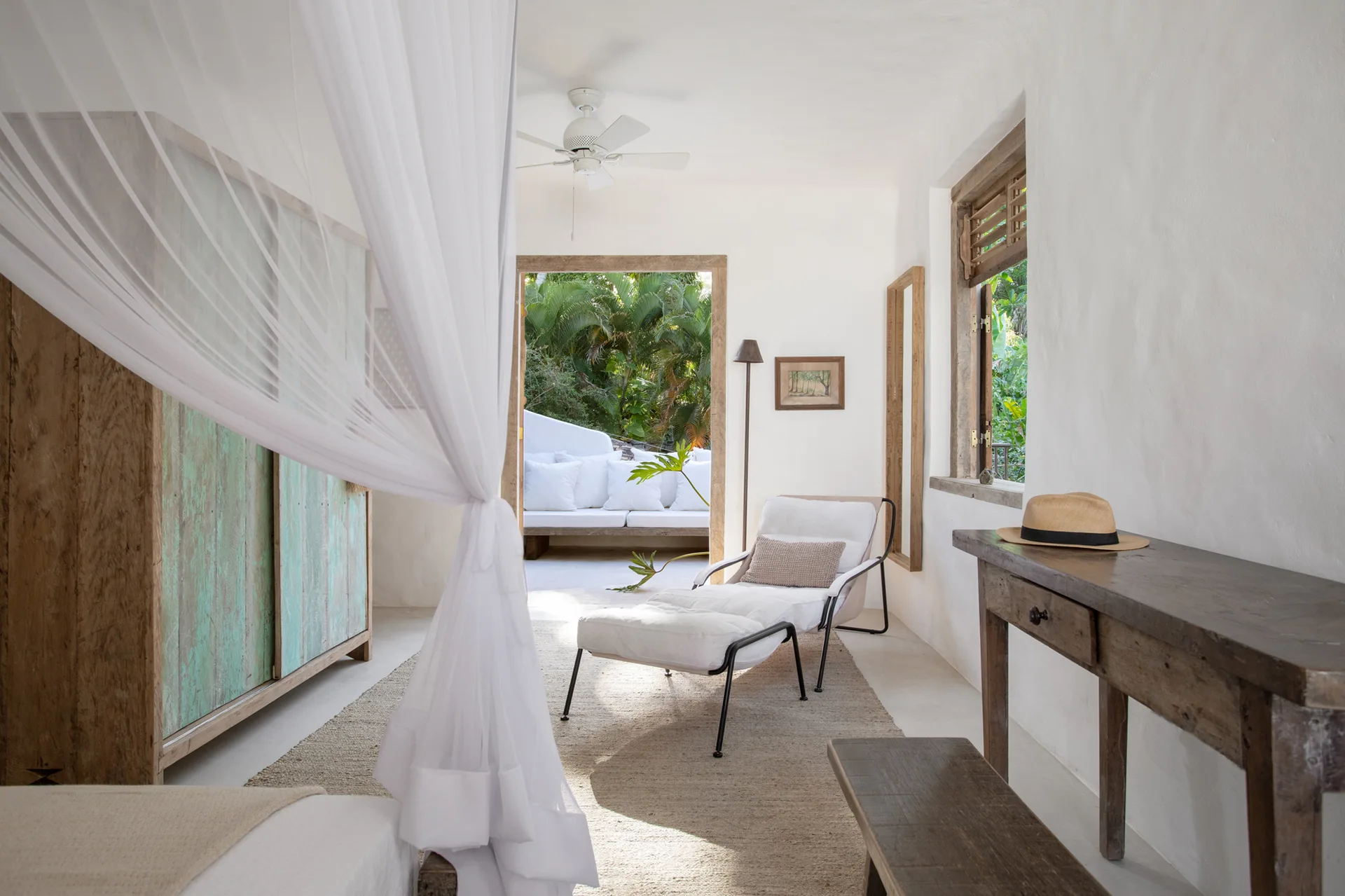 A bright white bedroom features a sheer bed canopy, a distressed wood wardrobe, and a white lounge chair facing an open doorway.