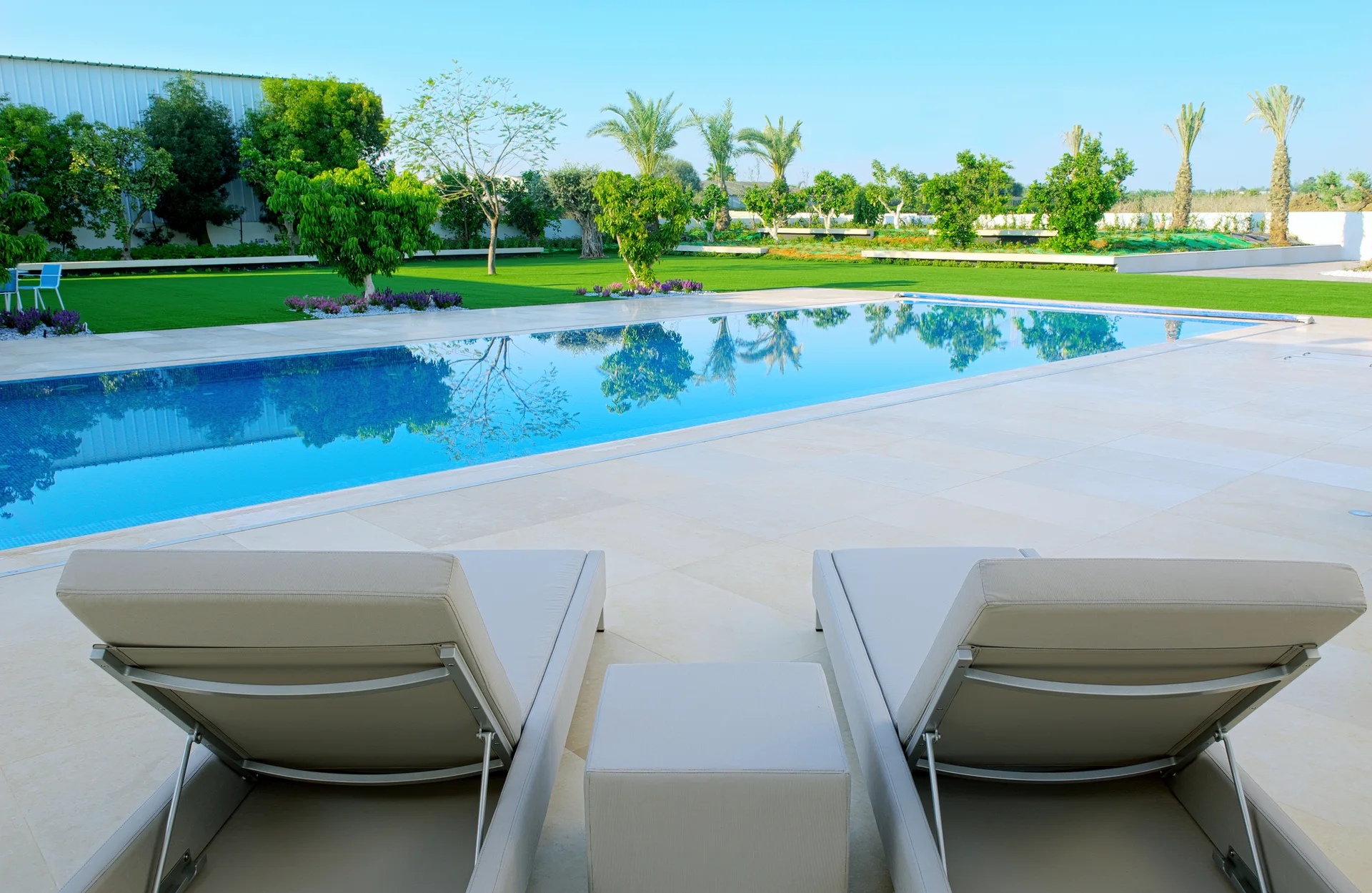 Two beige lounge chairs and a matching side table sit on a large-format stone tile patio next to a rectangular backyard swimming pool.