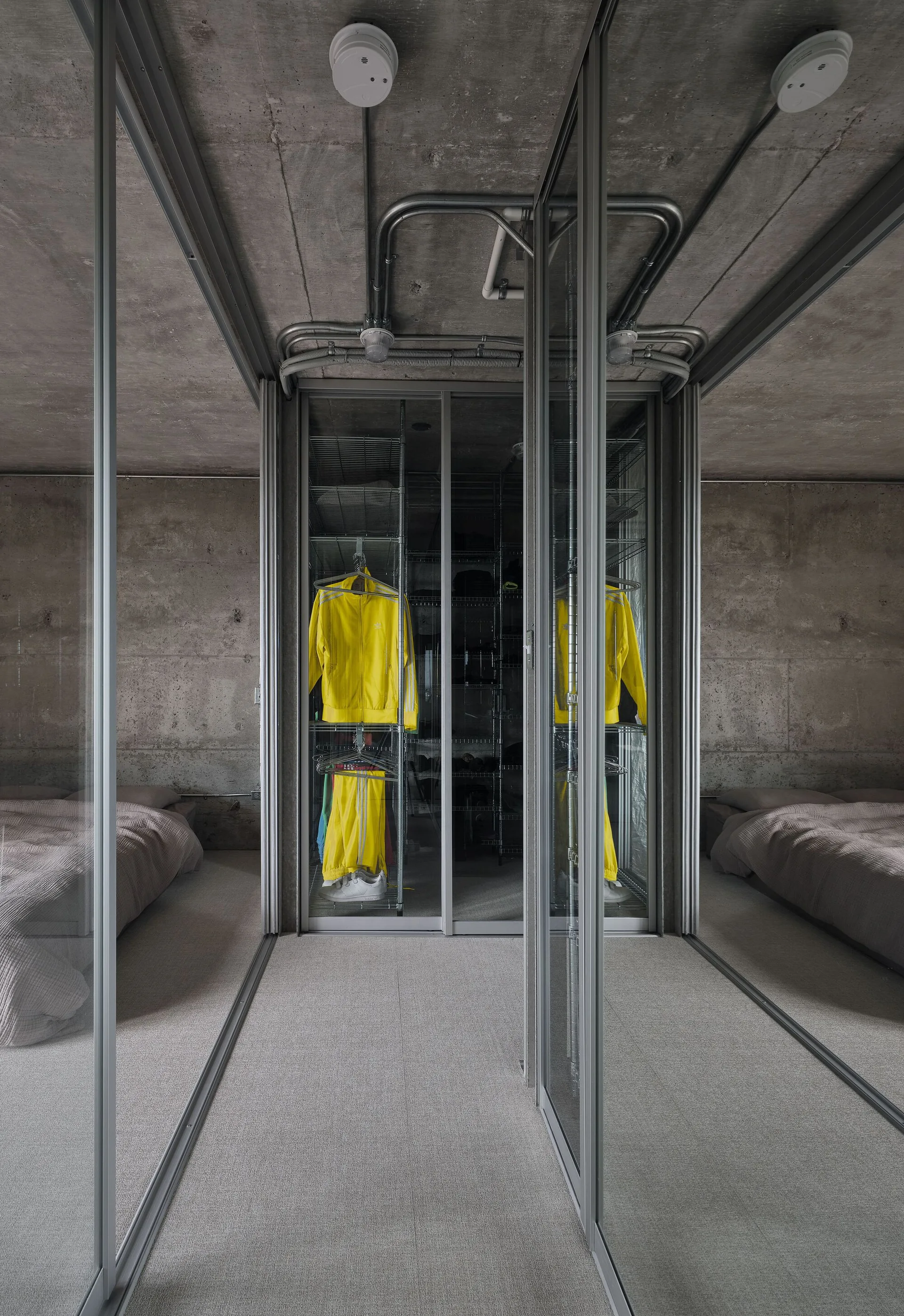 A modern industrial bedroom with exposed concrete walls, a concrete ceiling with metal conduit, and a glass-enclosed wardrobe.