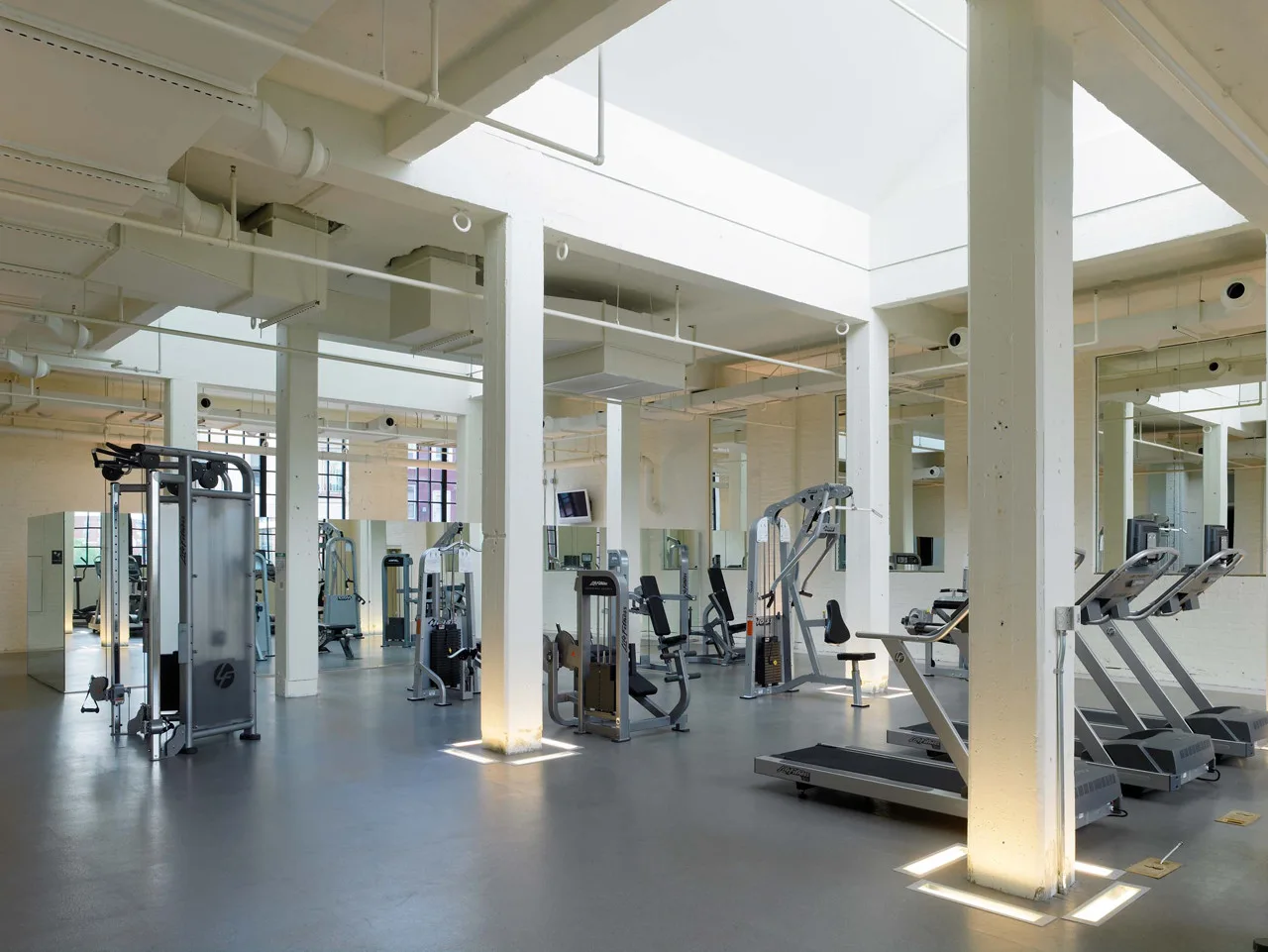 An industrial fitness center features exercise equipment, white structural columns, a large skylight, and grey flooring.