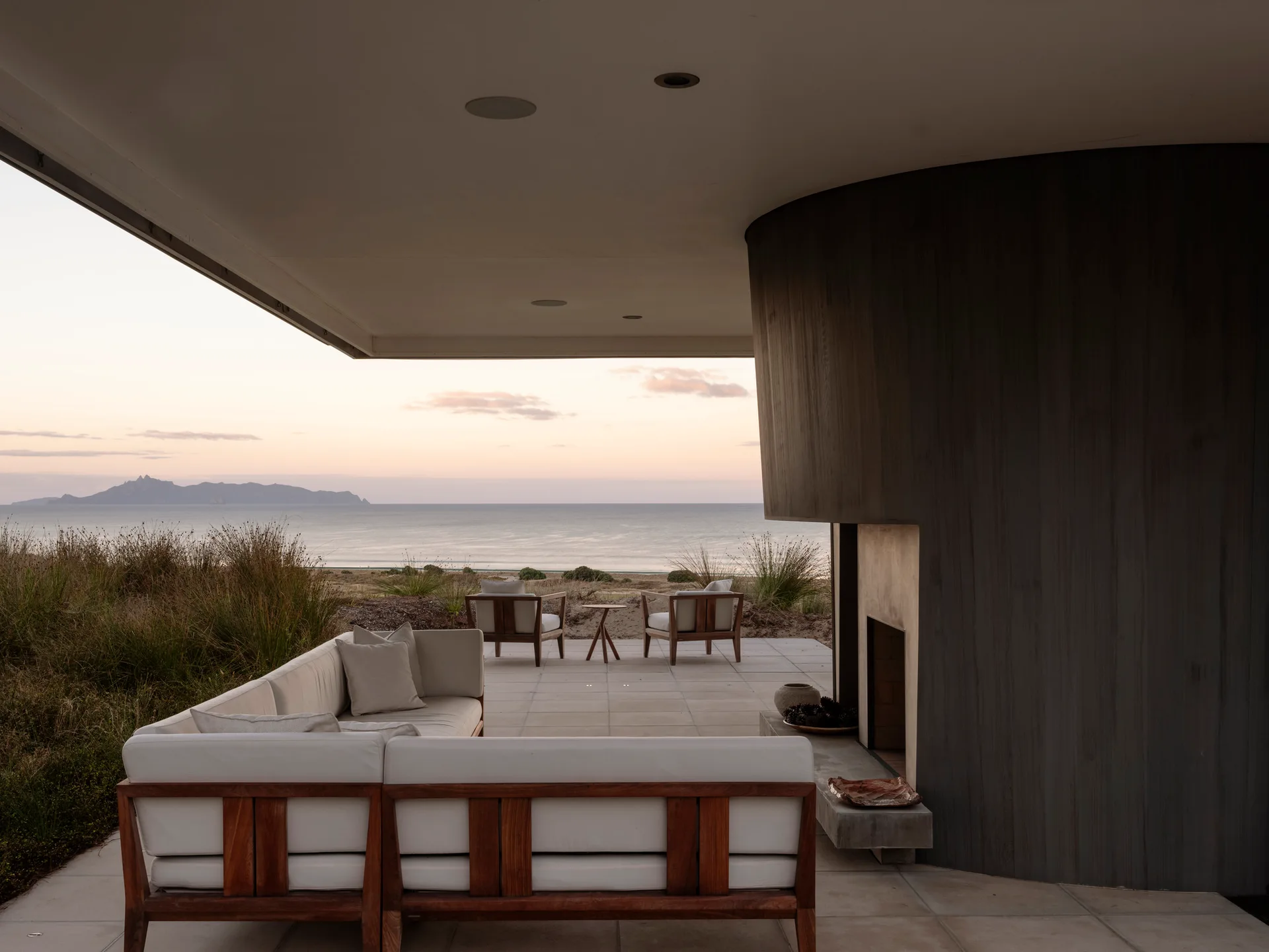 A covered concrete patio features modern wood-framed white sofas, a curved charcoal fireplace, and a clear view of the ocean at sunset.