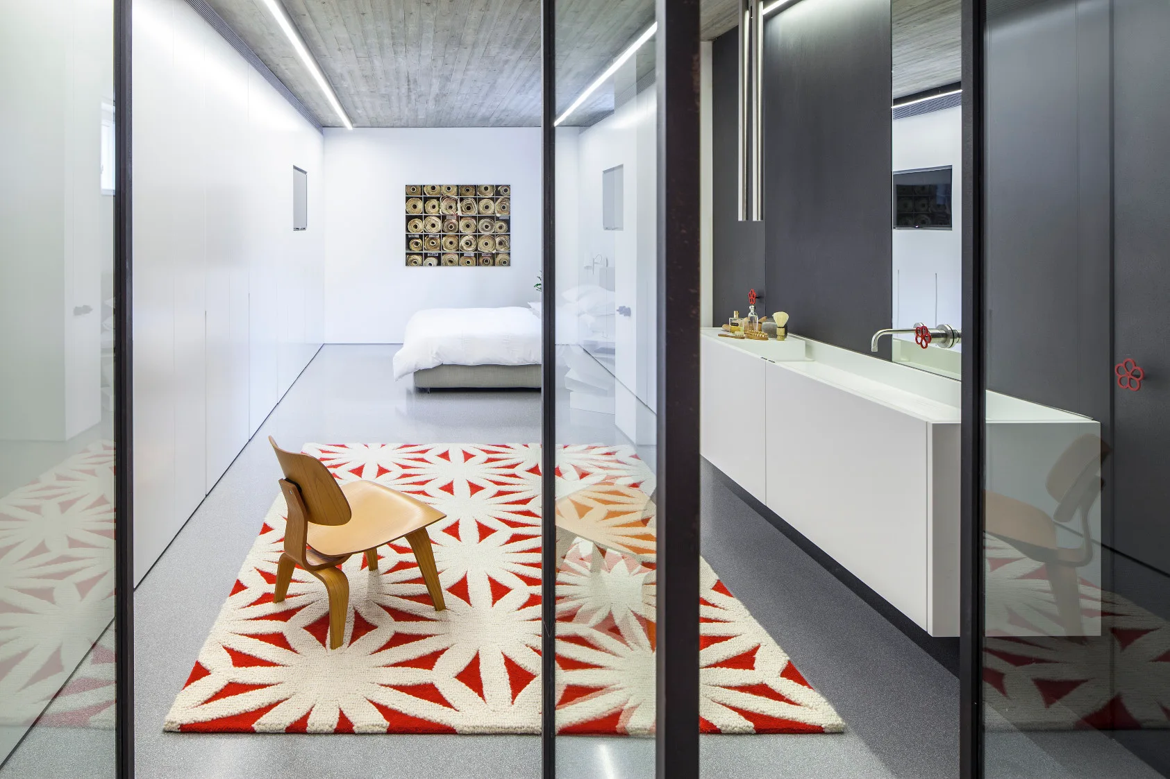 A modern bedroom and vanity suite features glass partitions, a concrete ceiling, a molded plywood chair on a red and white geometric rug, and white bedding.