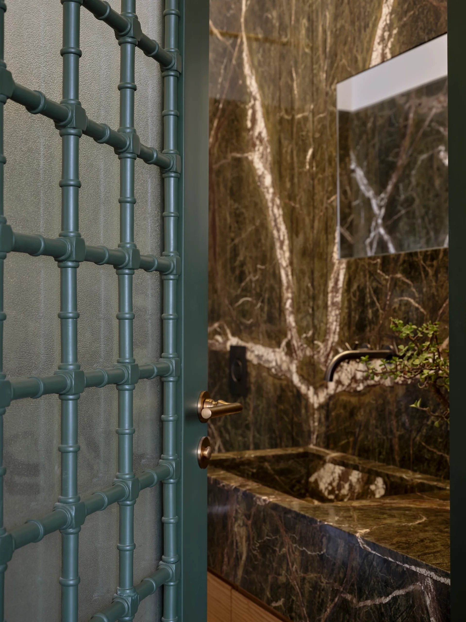 A green gridded glass door opens to a bathroom vanity with a matching dark green marble sink and marble-paneled wall.