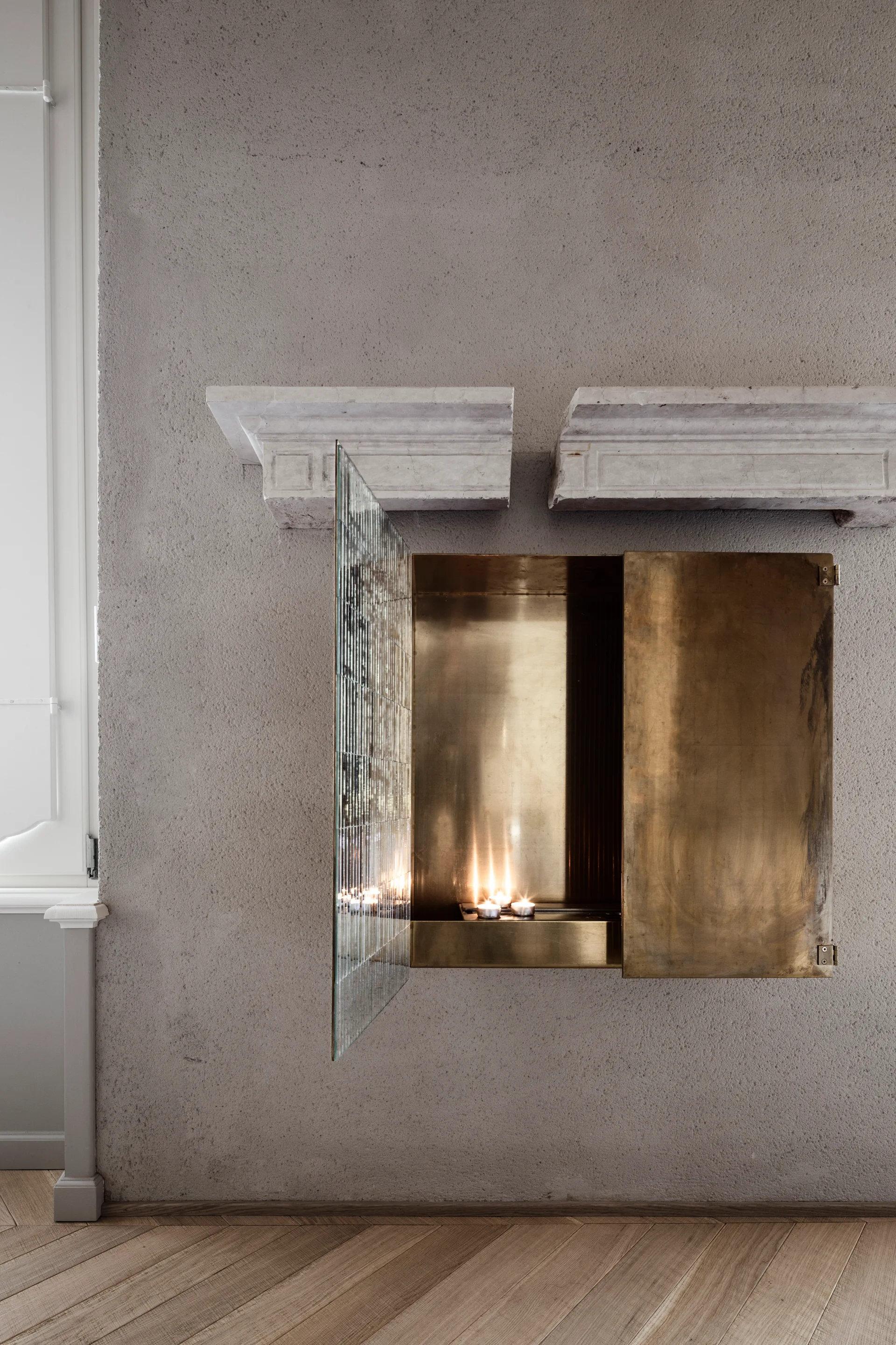 A recessed brass fireplace niche with lit candles and an open glass door is set into a textured plaster wall below a split stone mantel.