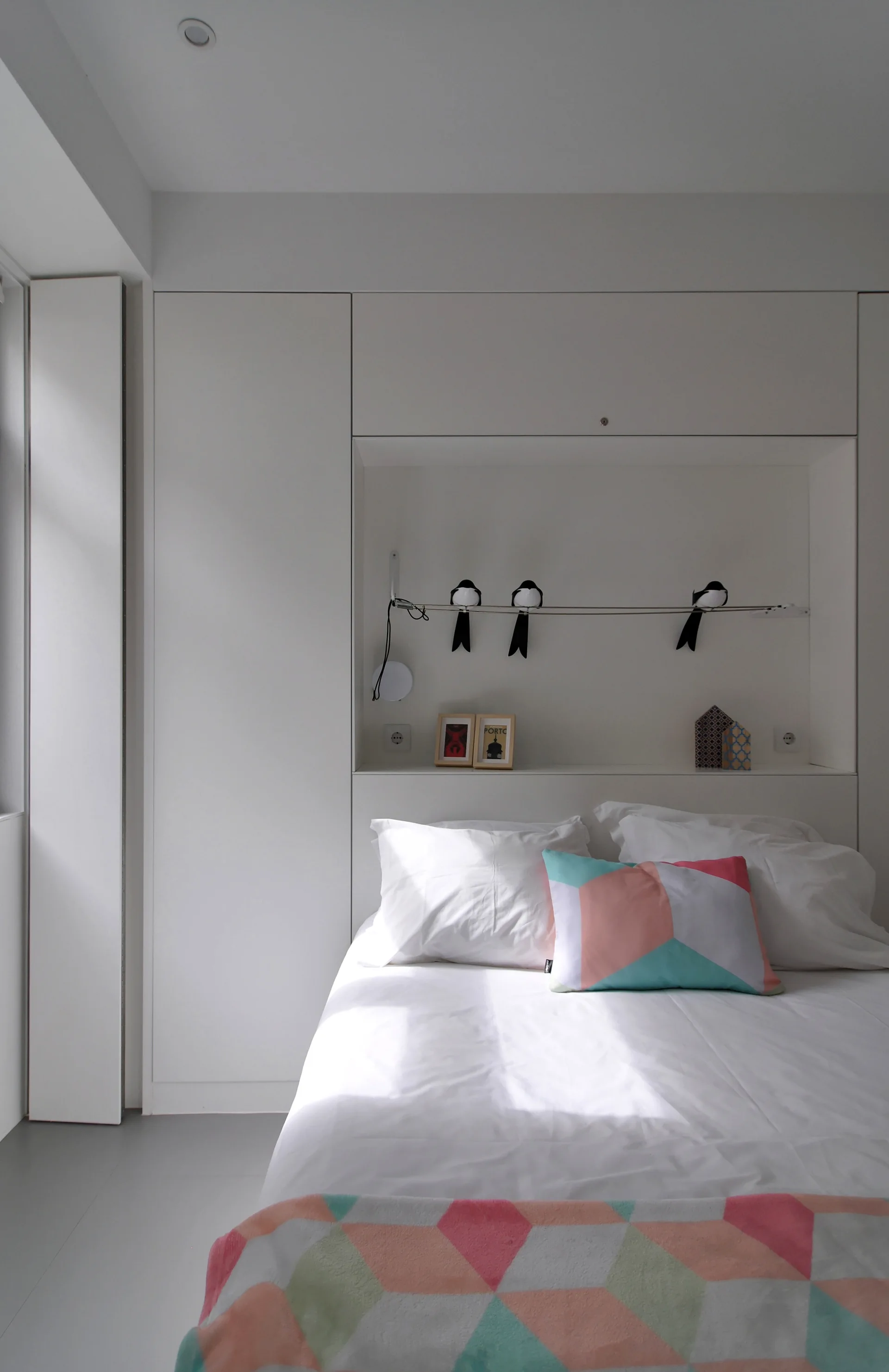 A minimalist white bedroom features a bed with geometric-patterned pillows set against integrated cabinetry with a built-in display niche.