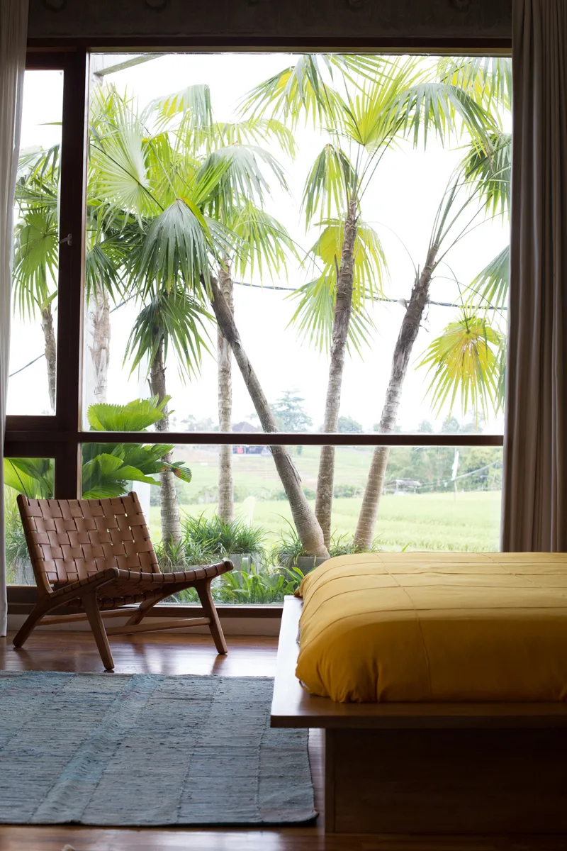 A low wooden platform bed with mustard yellow bedding sits next to a woven leather lounge chair in front of a large window framing green palm trees.
