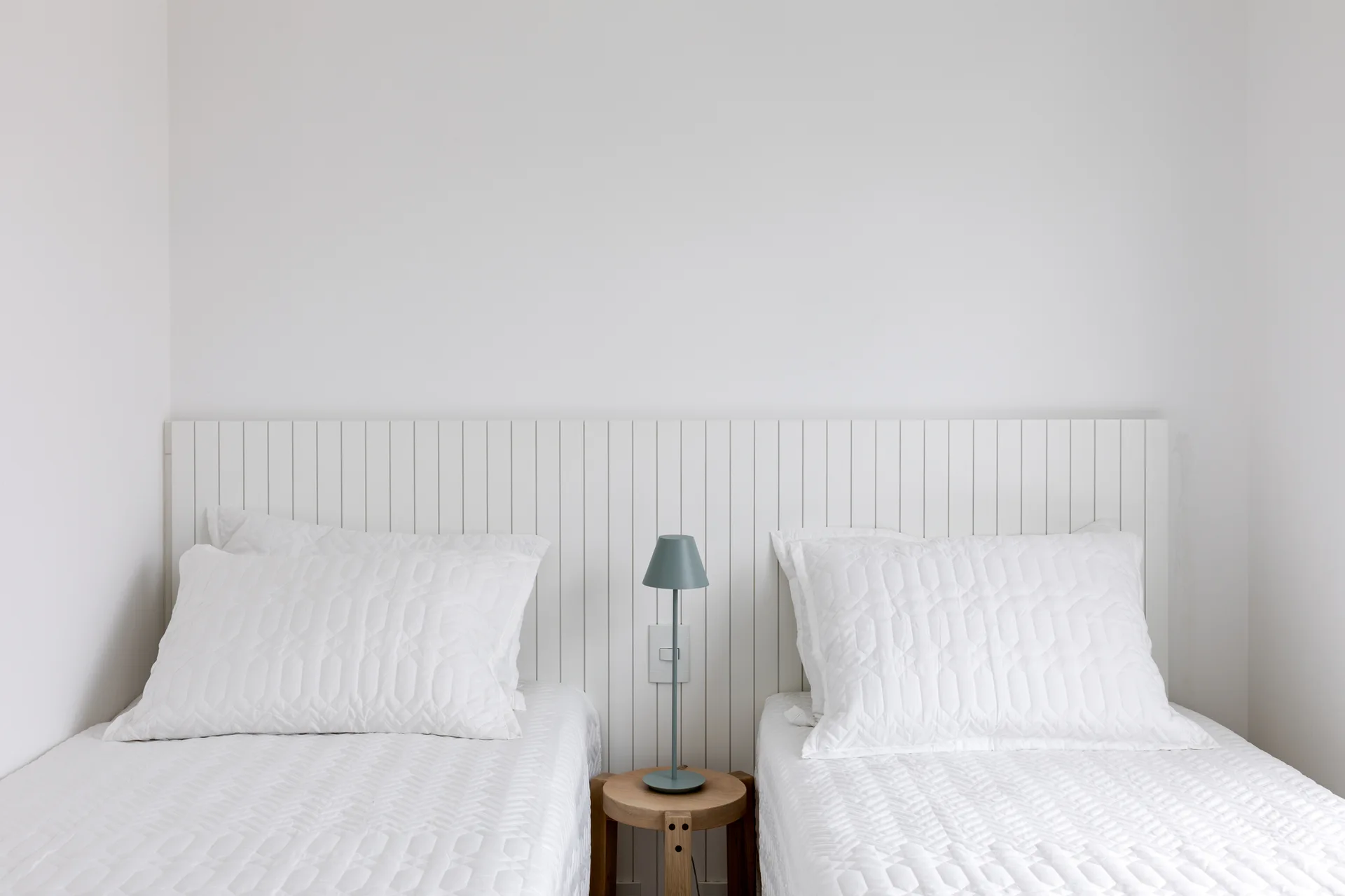 Two twin beds with textured white bedding sit side by side against a white vertical wood slat headboard, separated by a small round side table.