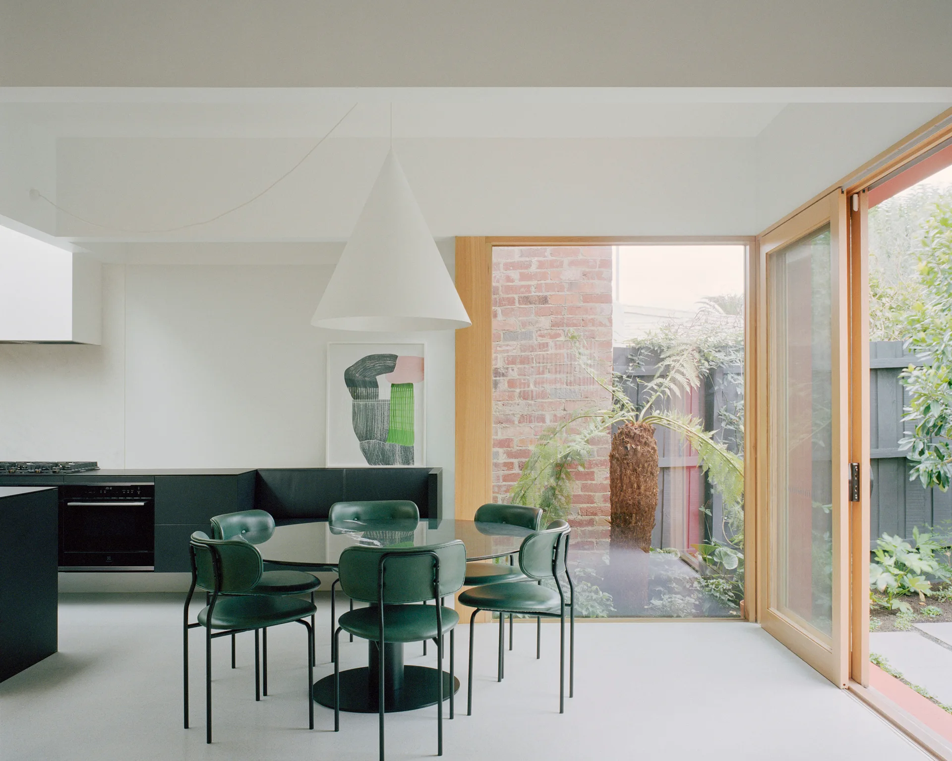 A minimalist dining area features a round glass table with forest green chairs, a white cone pendant light, and a large window showing a brick garden.