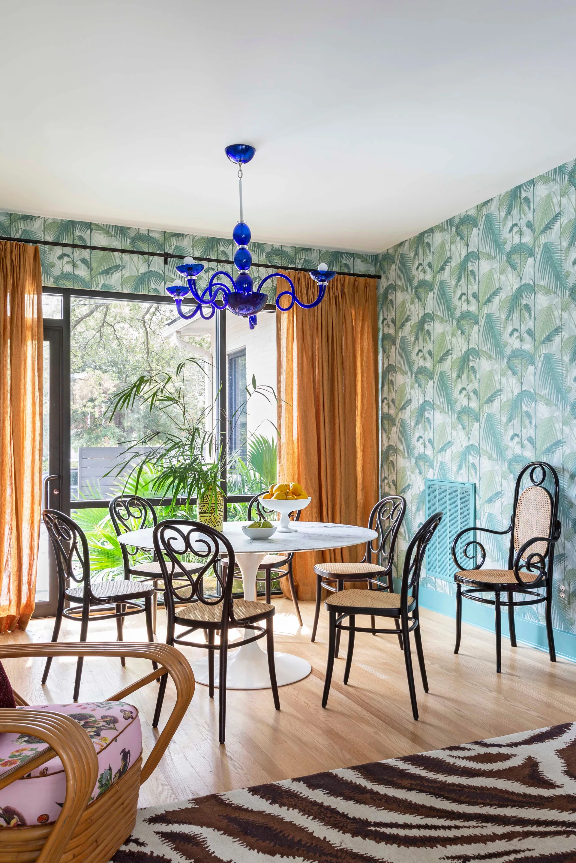 An eclectic dining room featuring a white round marble pedestal table, black bentwood chairs with rattan seats, palm leaf wallpaper, and a blue chandelier.