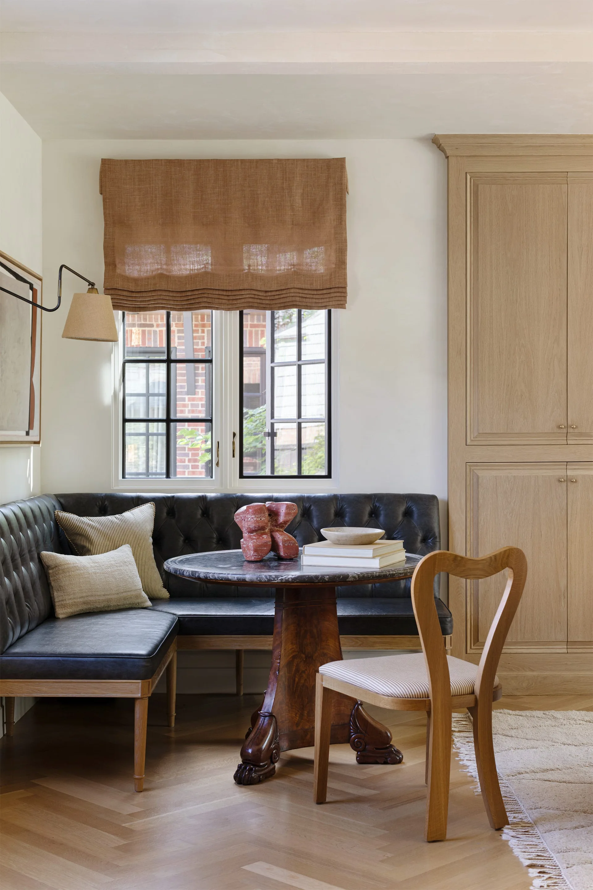 A breakfast nook features a black tufted leather corner banquette, a round stone pedestal table, a wooden chair, and tall oak cabinets.