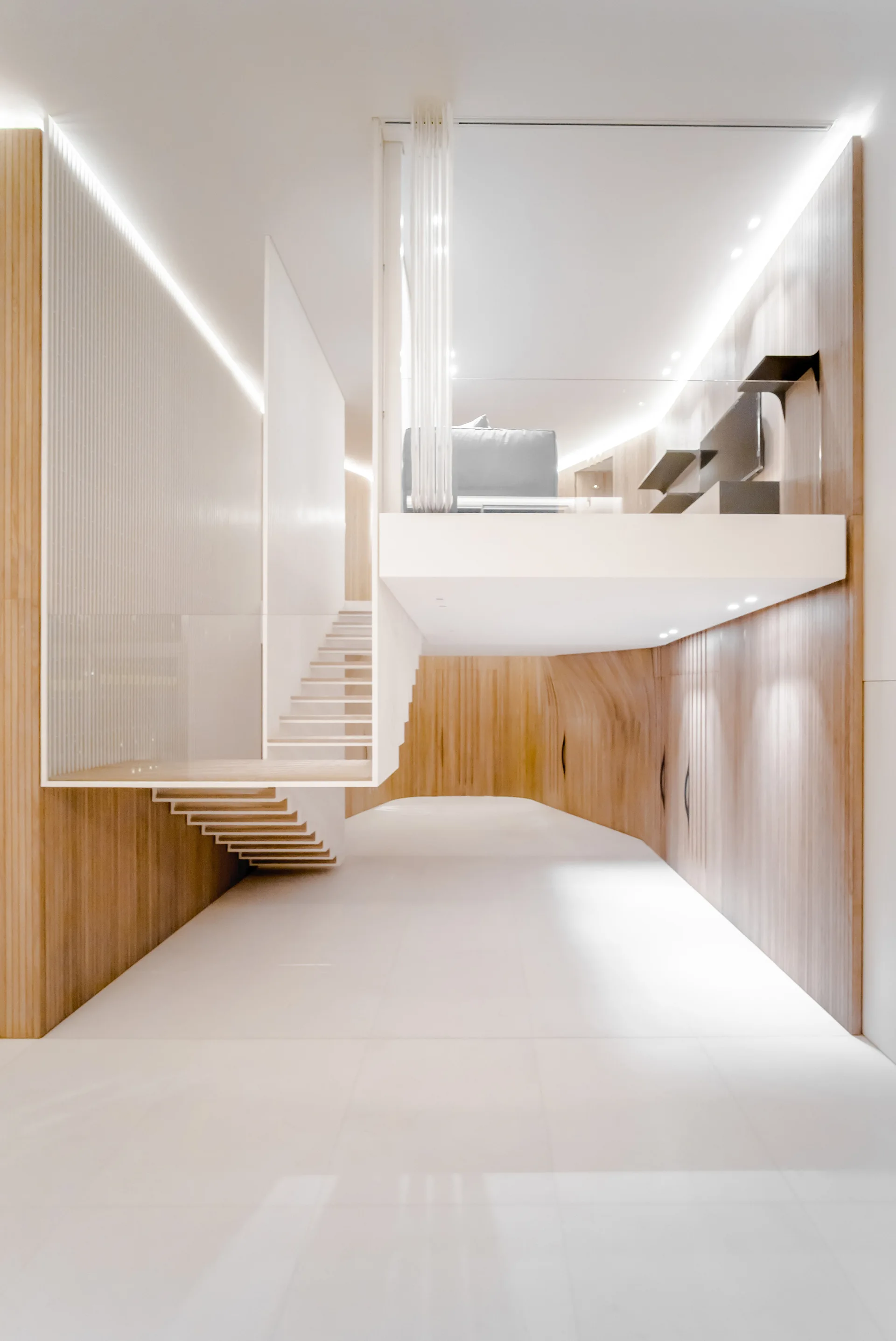 A minimalist interior featuring a white floating staircase, wood-paneled walls, and an elevated mezzanine level with integrated cove lighting.