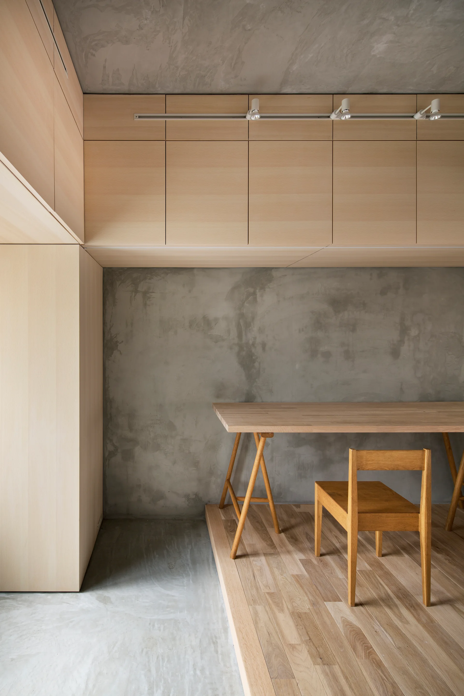 A minimalist home office workspace features a light wood desk and chair on a raised wooden platform against grey concrete walls with overhead cabinets.