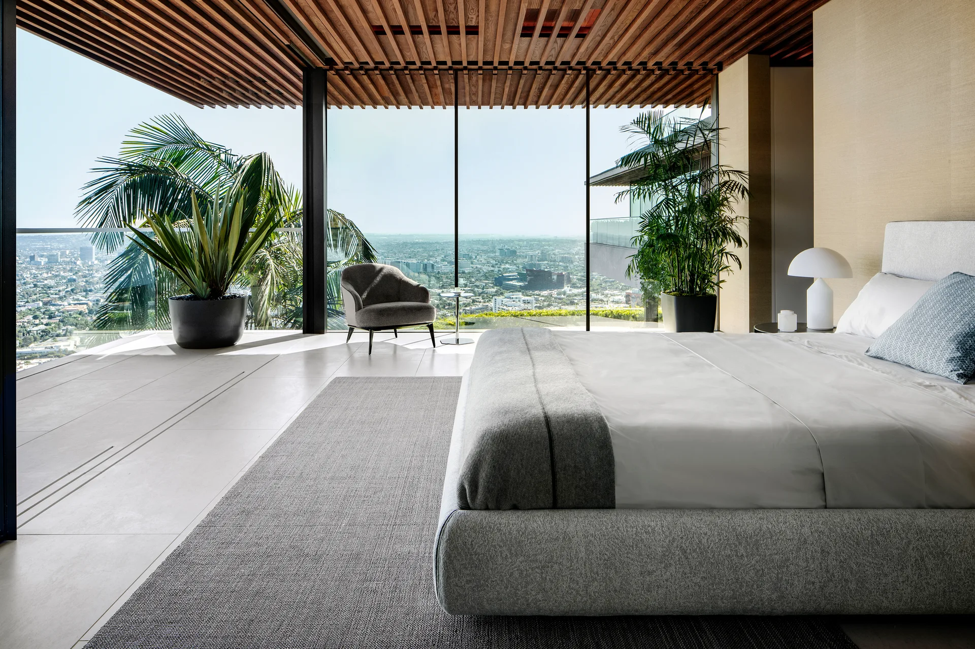 A modern bedroom features a grey upholstered bed, a slatted wood ceiling, and floor-to-ceiling glass walls looking out over a city.