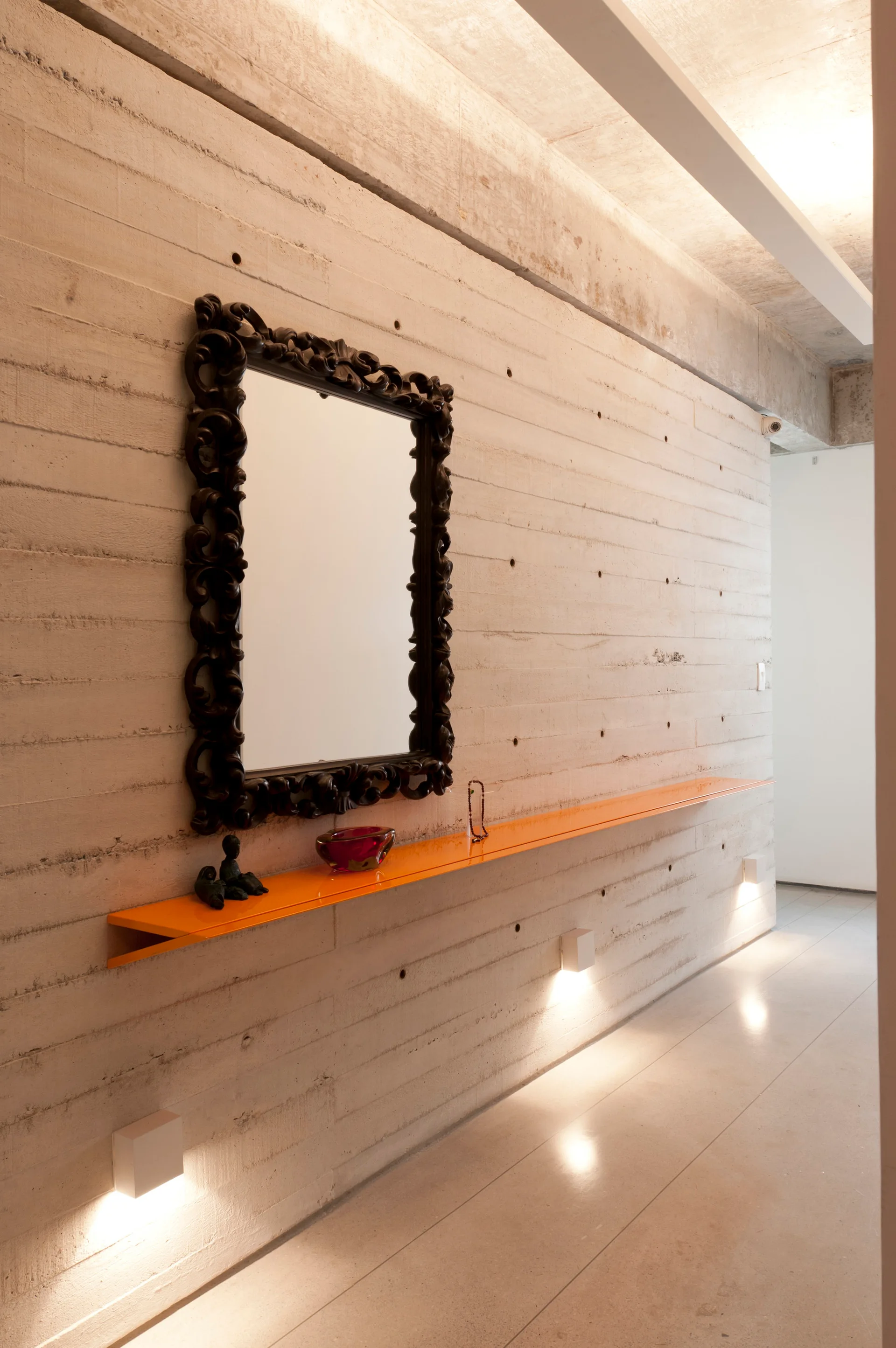 A minimalist hallway features a board-formed concrete wall, a long bright orange floating shelf, and a dark ornate carved mirror.