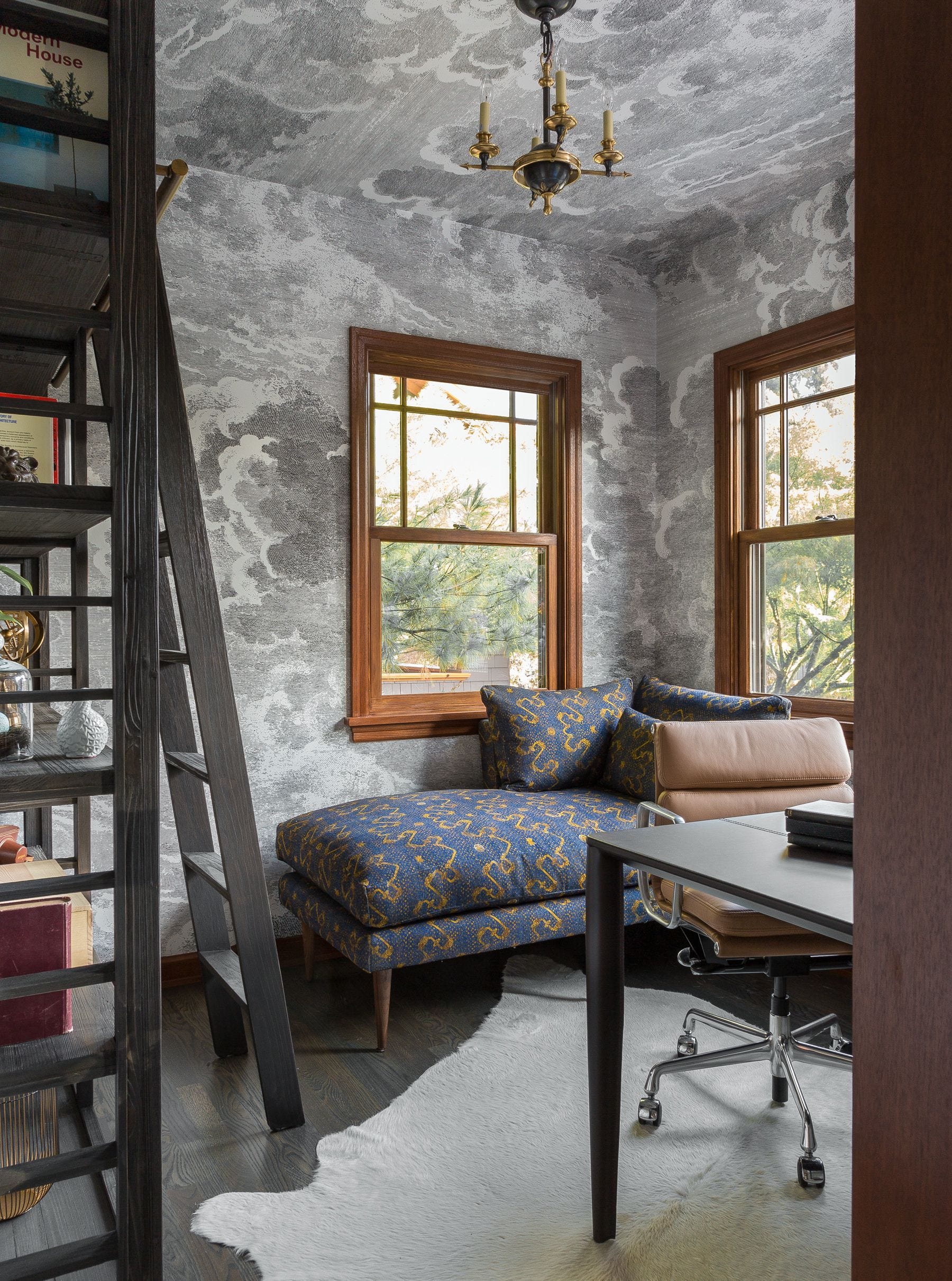 A home office features gray cloud-patterned wallpaper on the walls and ceiling, a blue chaise lounge, and a dark wood bookshelf with a ladder.