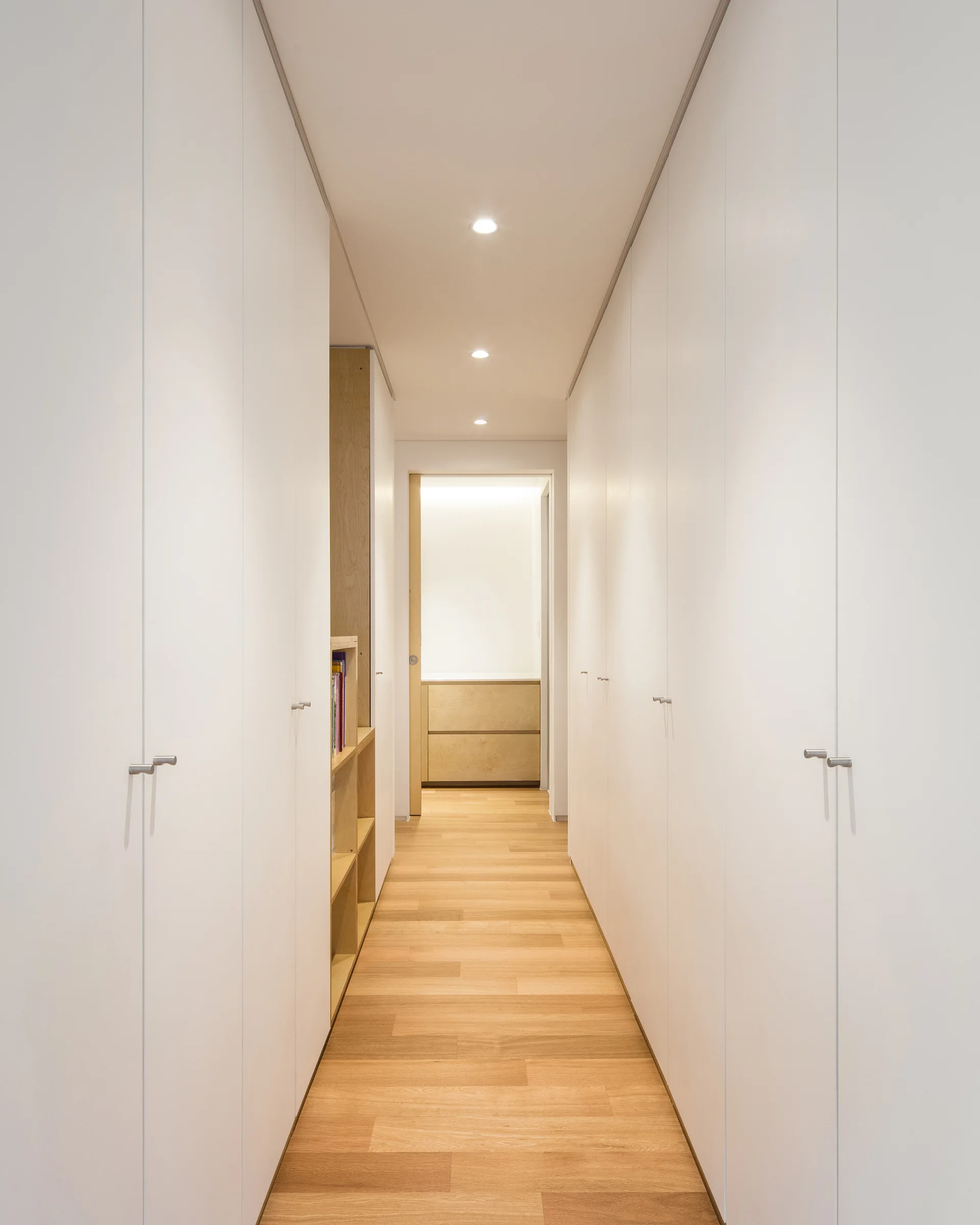 A narrow residential hallway featuring floor-to-ceiling white built-in closets with small silver handles and light oak wood flooring.