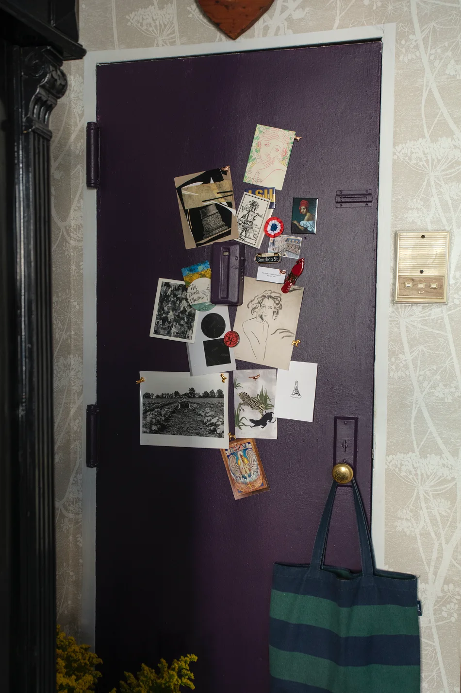 A deep purple entryway door pinned with postcards and small art prints, set against beige floral wallpaper with a striped tote bag on the brass knob.