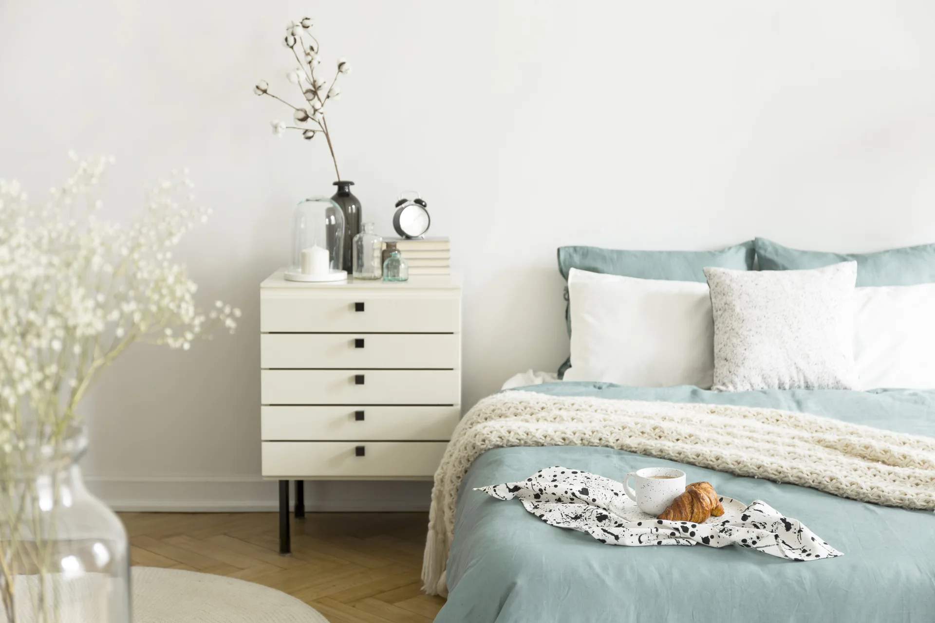A minimalist bedroom featuring sage green linen bedding, a white five-drawer dresser as a nightstand, and light herringbone floors.