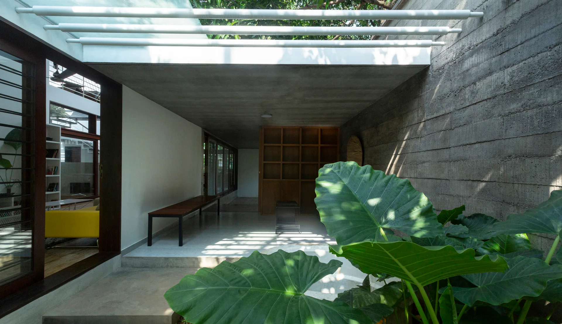 A transitional courtyard features a textured concrete wall, a wooden bench, a built-in bookshelf, and large green tropical leaves in the foreground.